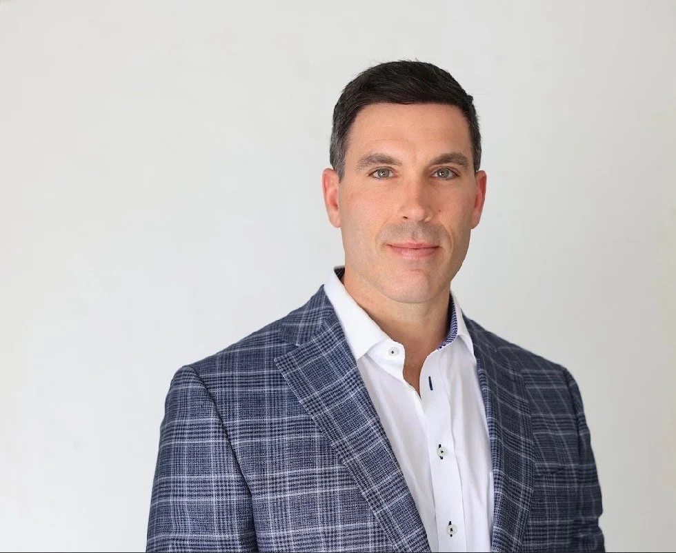 Headshot of a man with short dark hair, blue eyes, wearing a checkered blazer and white shirt, against a plain white background.