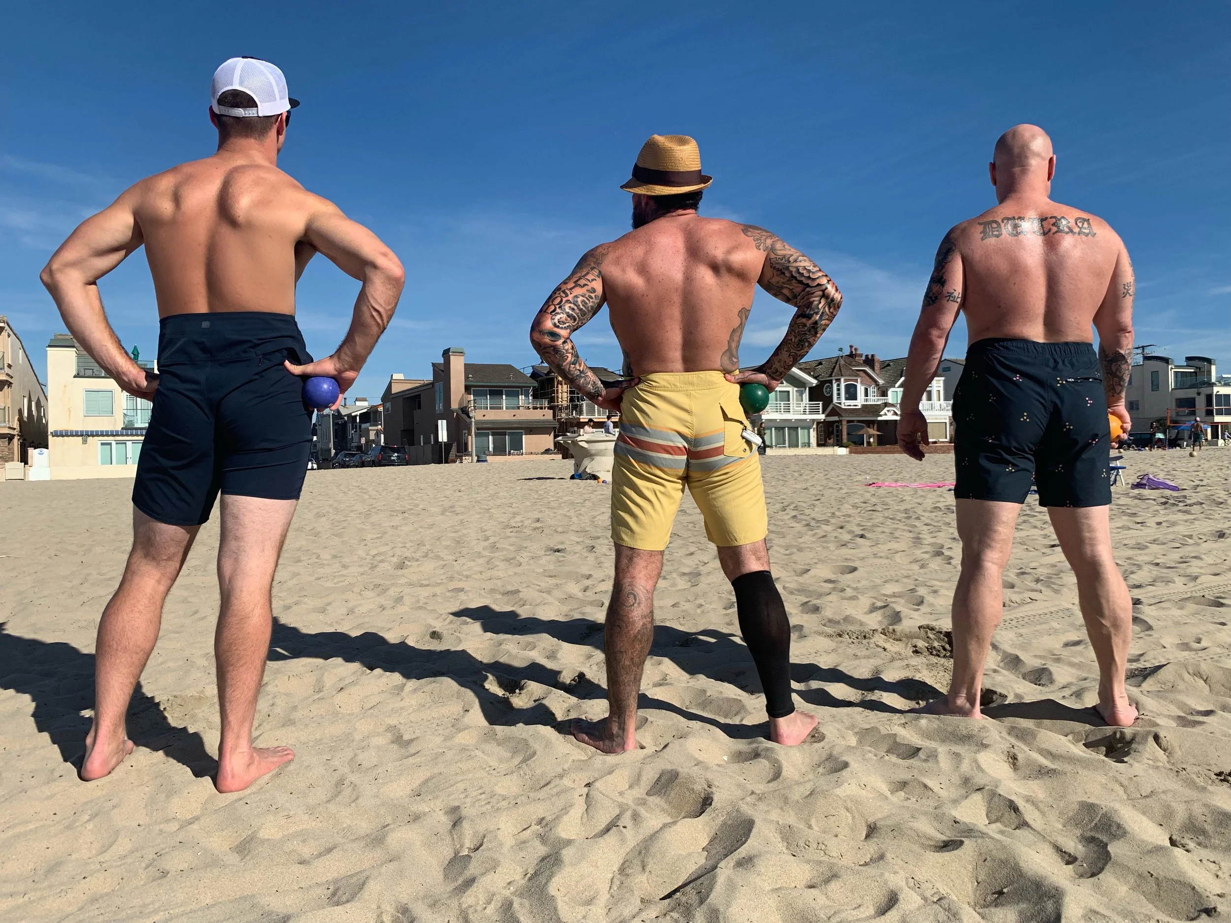 Three men are standing on a sandy beach with their backs to the camera, facing the ocean under a clear blue sky. The man on the left is wearing a white golf cap and black swim trunks, the man in the middle has a straw hat and yellow swim trunks, and 