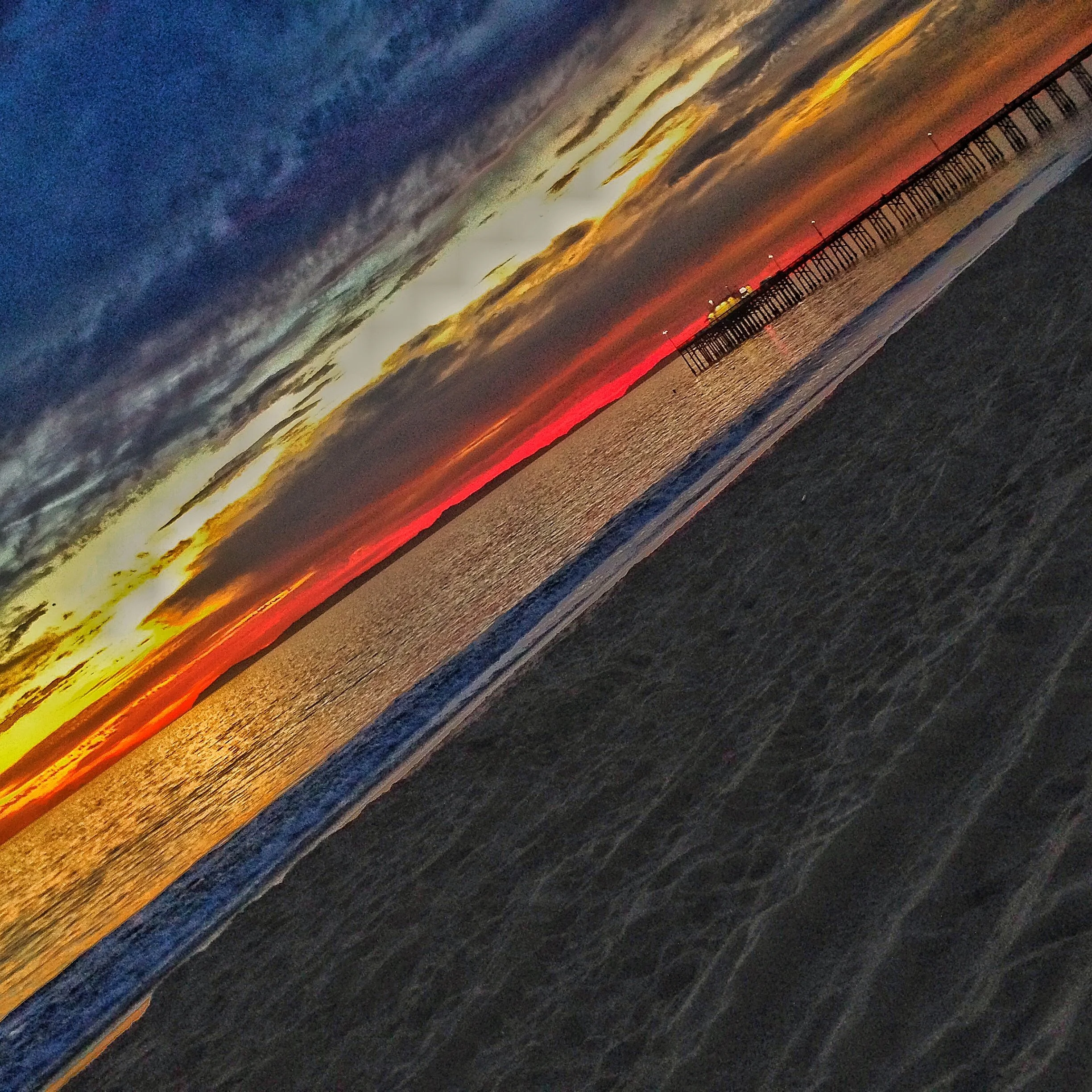 Sunset over the ocean with the balboa pier extending into the water, colorful clouds in the sky, and reflections on the wet sand.