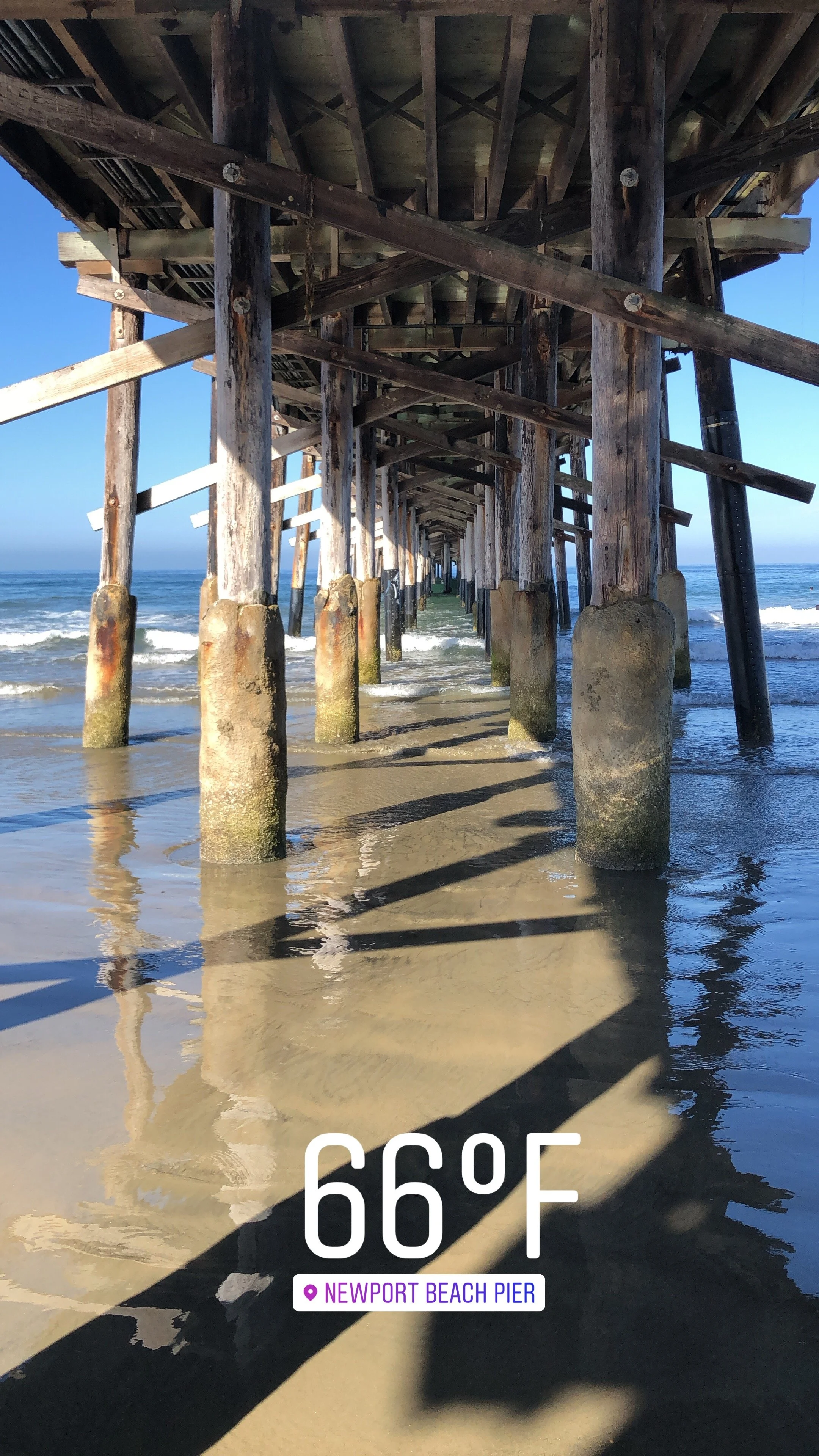 View of a wooden pier extending into the ocean, with waves hitting the supports. The weather is clear and sunny, and the temperature displayed is 66°F. The location indicated is Newport Beach Pier.