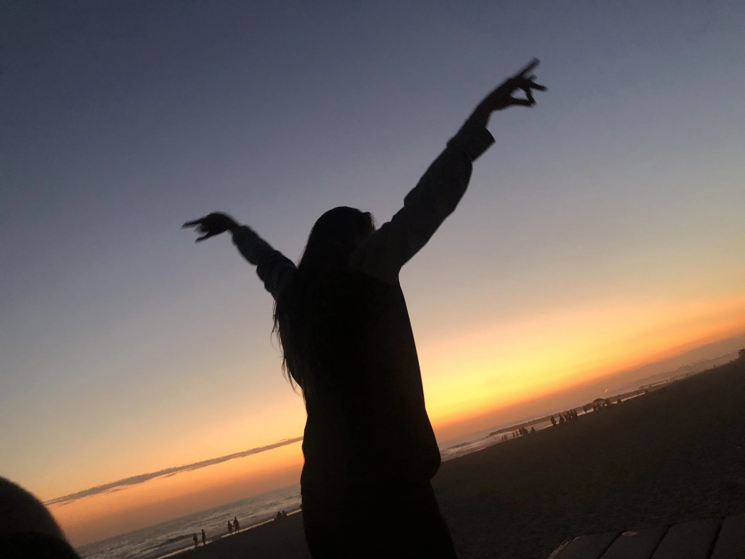 A person with long hair and wearing a dark jacket standing on a beach at sunset, raising their arms in a gesture with hands forming a triangle, with a silhouette of other people in the distance and a colorful sky.