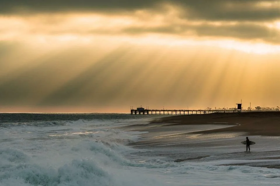 Beach scene at sunset with the newport beach pier extending into the ocean and a person carrying a surfboard along the shore.