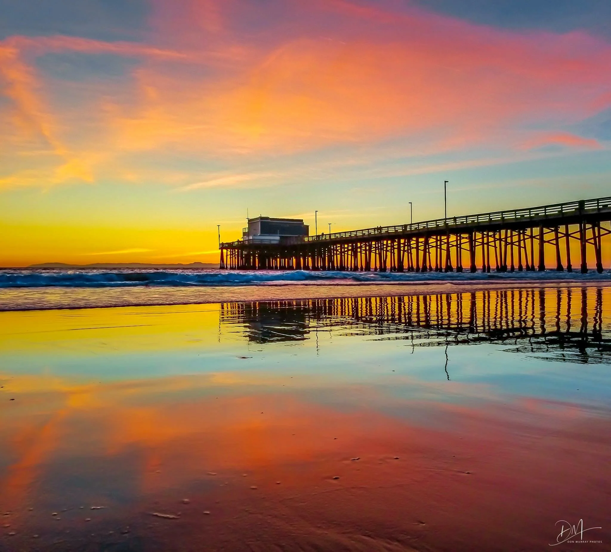 Sunset over the ocean with a pier extending into the water, colorful sky with pink, orange, and purple clouds, calm waves, and a small signature in the bottom right corner.