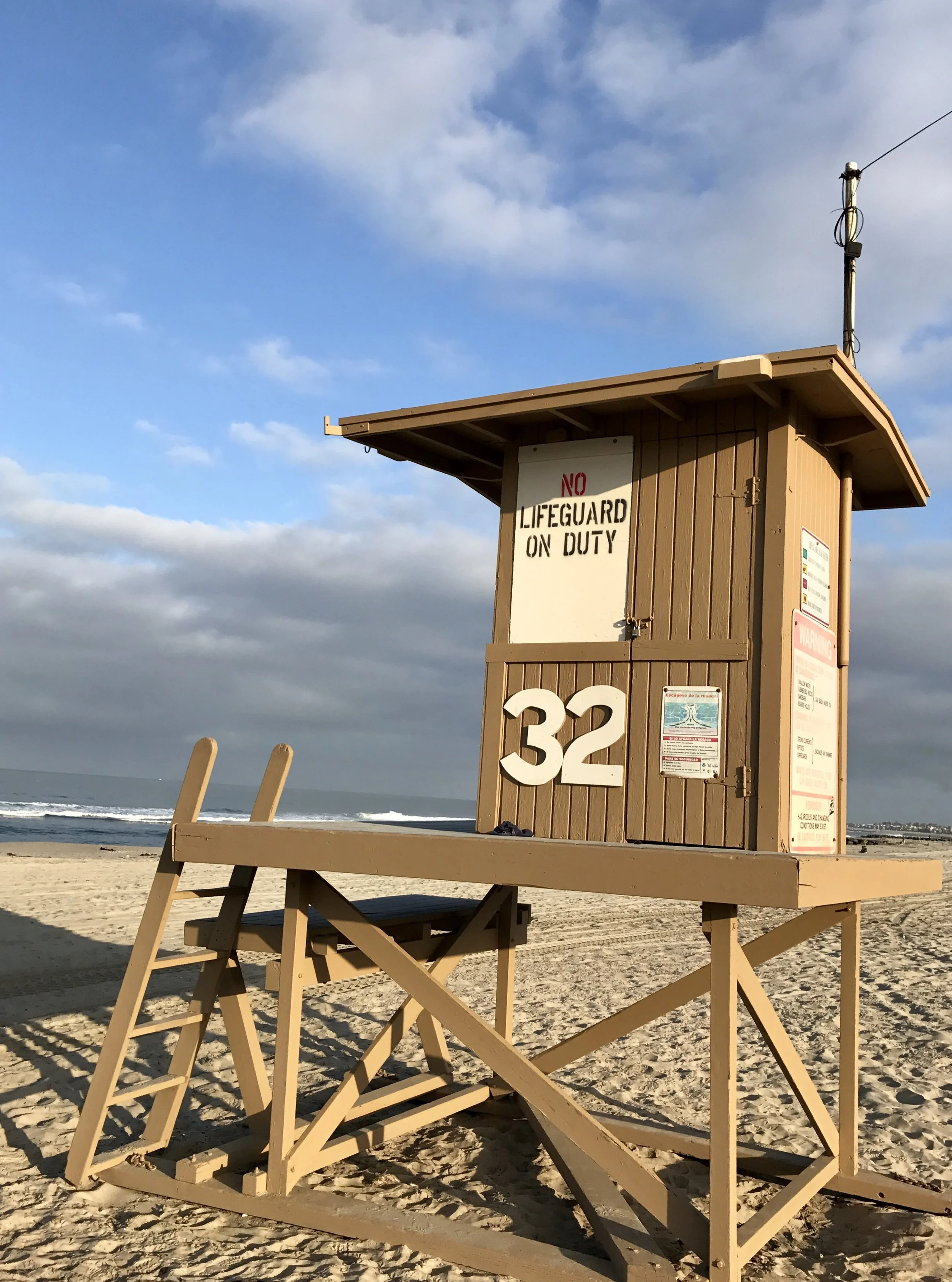 lifeguard tower number 32 on 32nd street in newport beach with a sign that reads "NO LIFEGUARD ON DUTY" and the number 32, against a backdrop of a blue sky with some clouds and ocean waves in the distance.