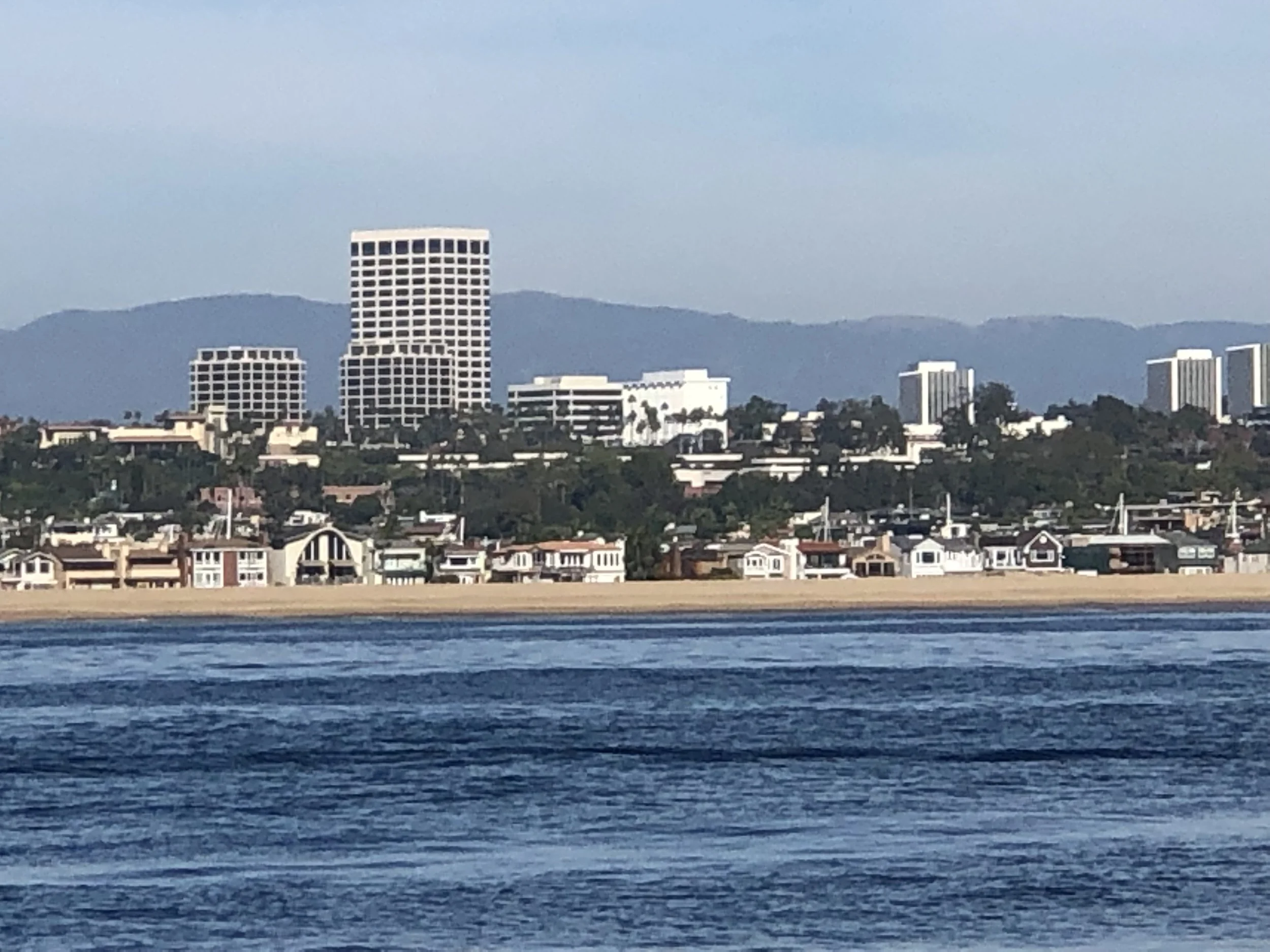 A city skyline with multiple high-rise buildings viewed across a body of water, with mountains in the background on a clear day.