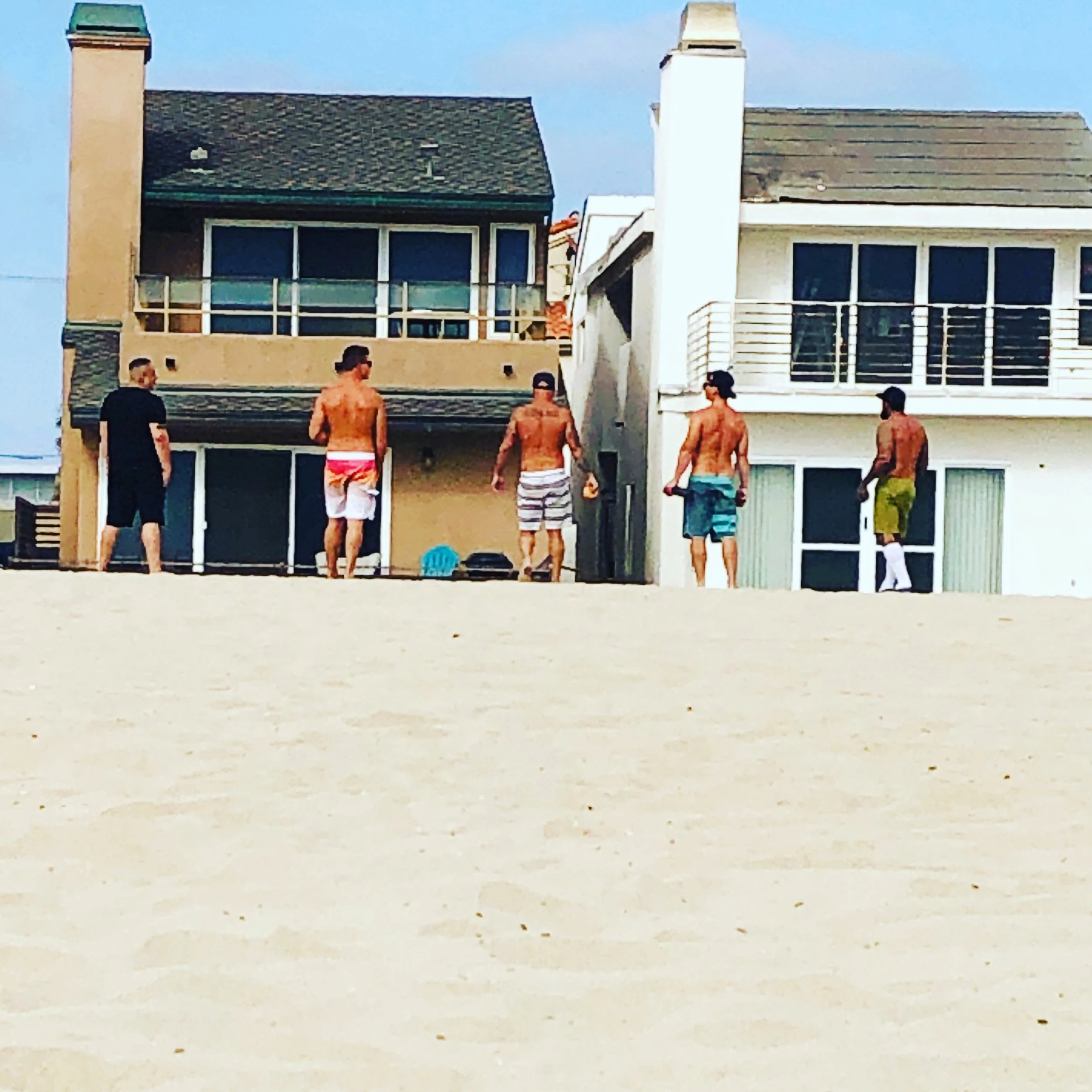Group of young men standing shirtless on a beach in front of residential buildings with balconies during daytime.