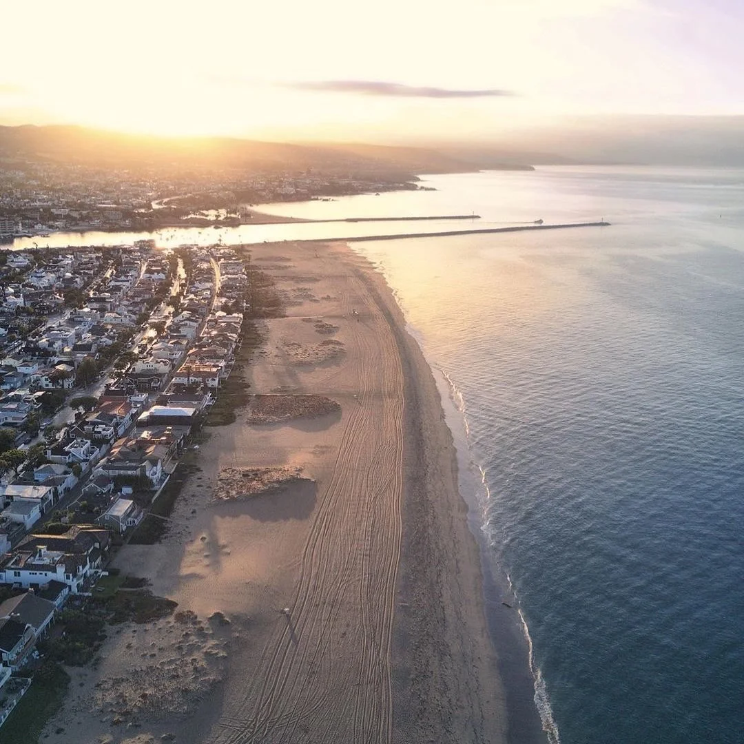Aerial view of a beach at sunset with residential houses on one side and ocean waves on the other, with a pier extending into the water in the background.