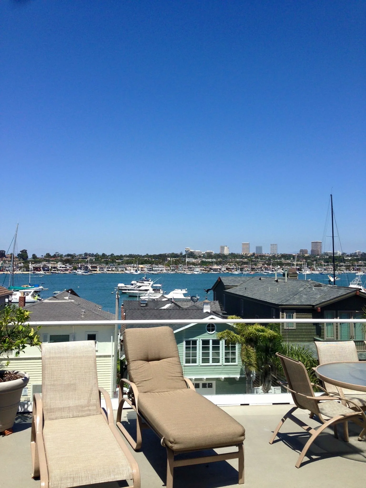 View of a balcony overlooking the newport harbor with numerous sailboats and yachts, with a city skyline and tall buildings in the background under a clear blue sky, and several lounge chairs and potted plants on the balcony.