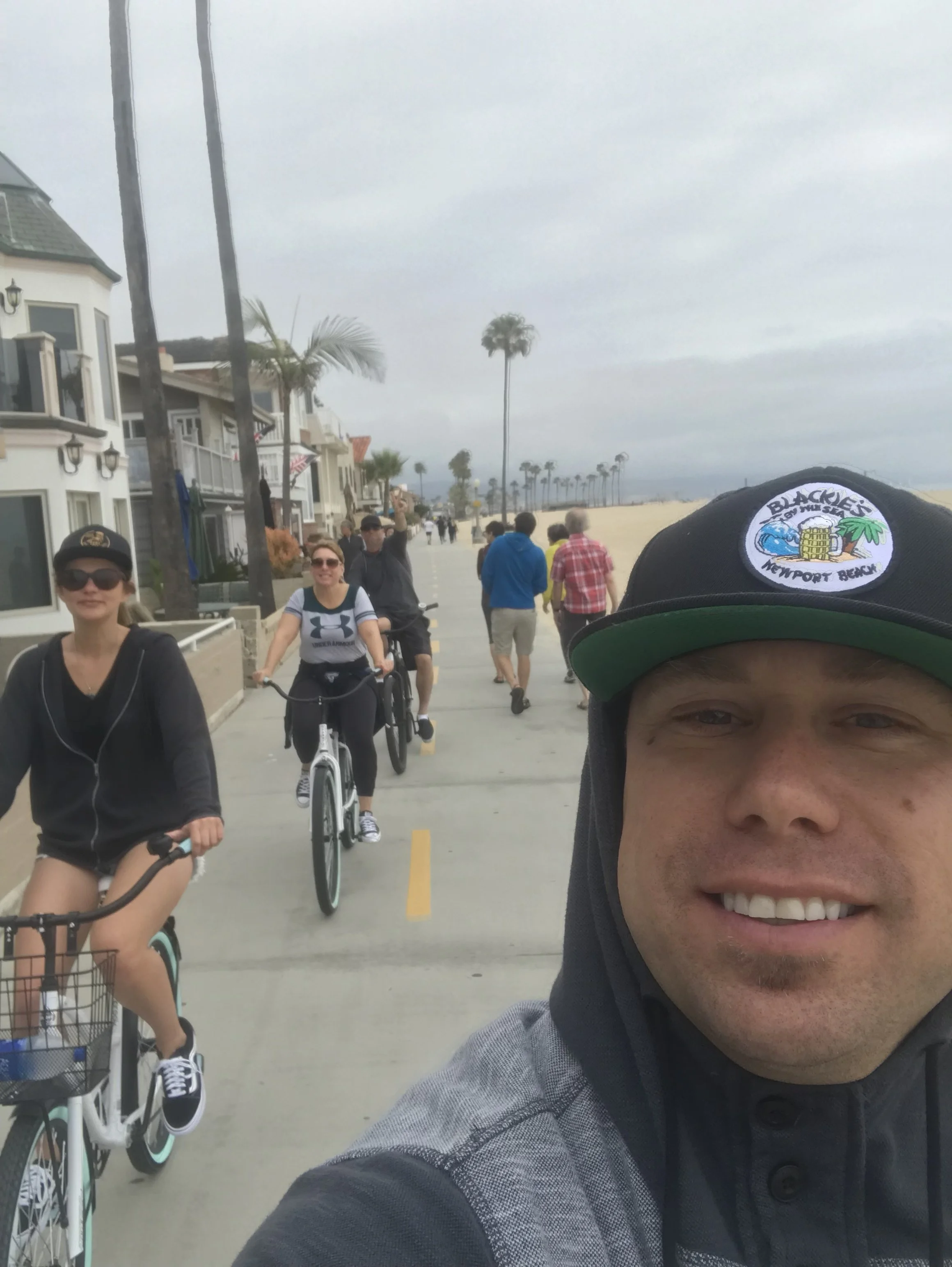 A man taking a selfie at the beach boardwalk with smiling women and people walking and biking, buildings, palm trees, and an overcast sky in the background.