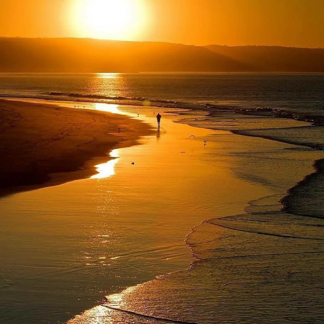 Sunset over a beach with a person walking along the shore, reflecting golden light, with hills in the background.