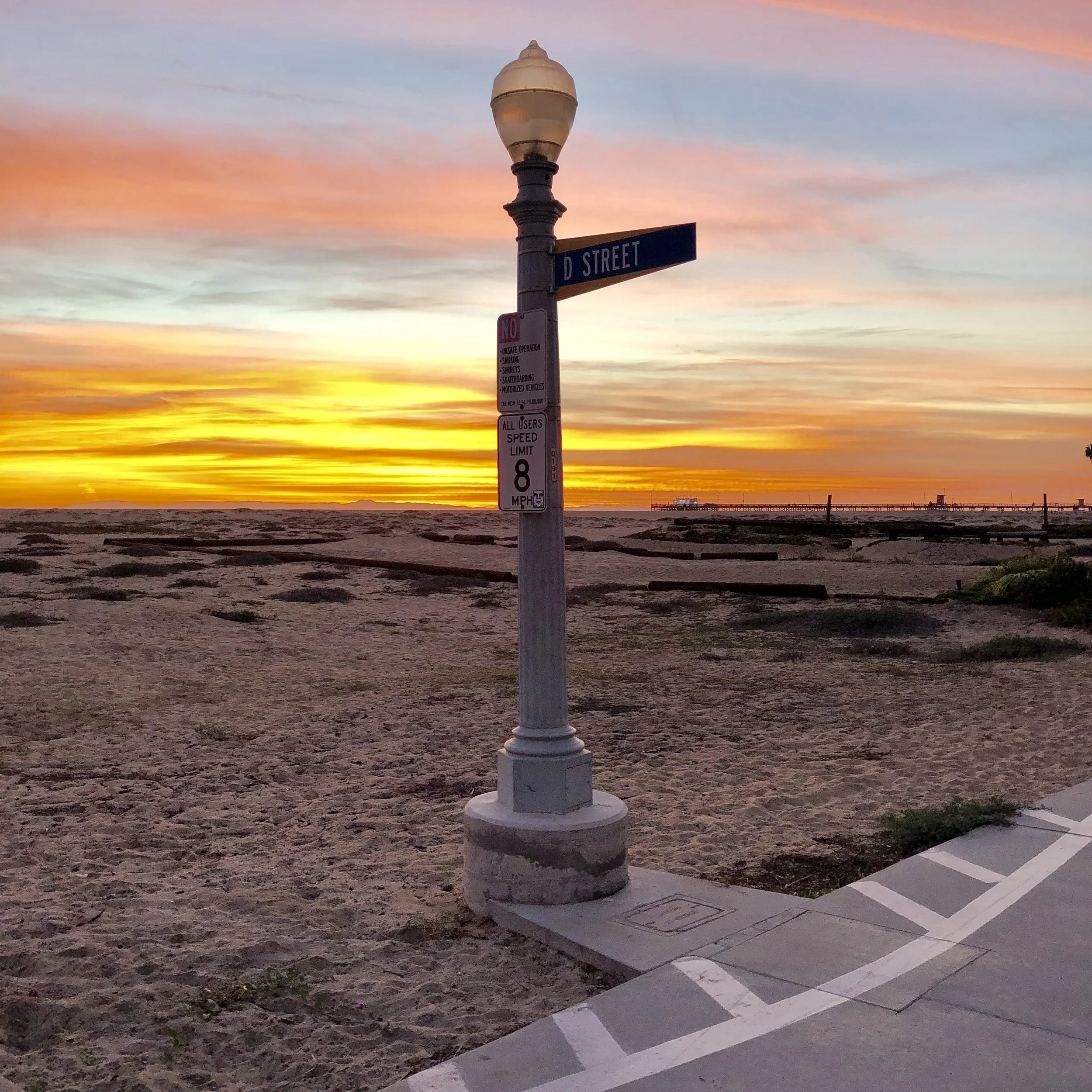 A streetlamp and a street sign on D Street during sunset with colorful pink, purple, and orange clouds in the sky, near a pier and the ocean.