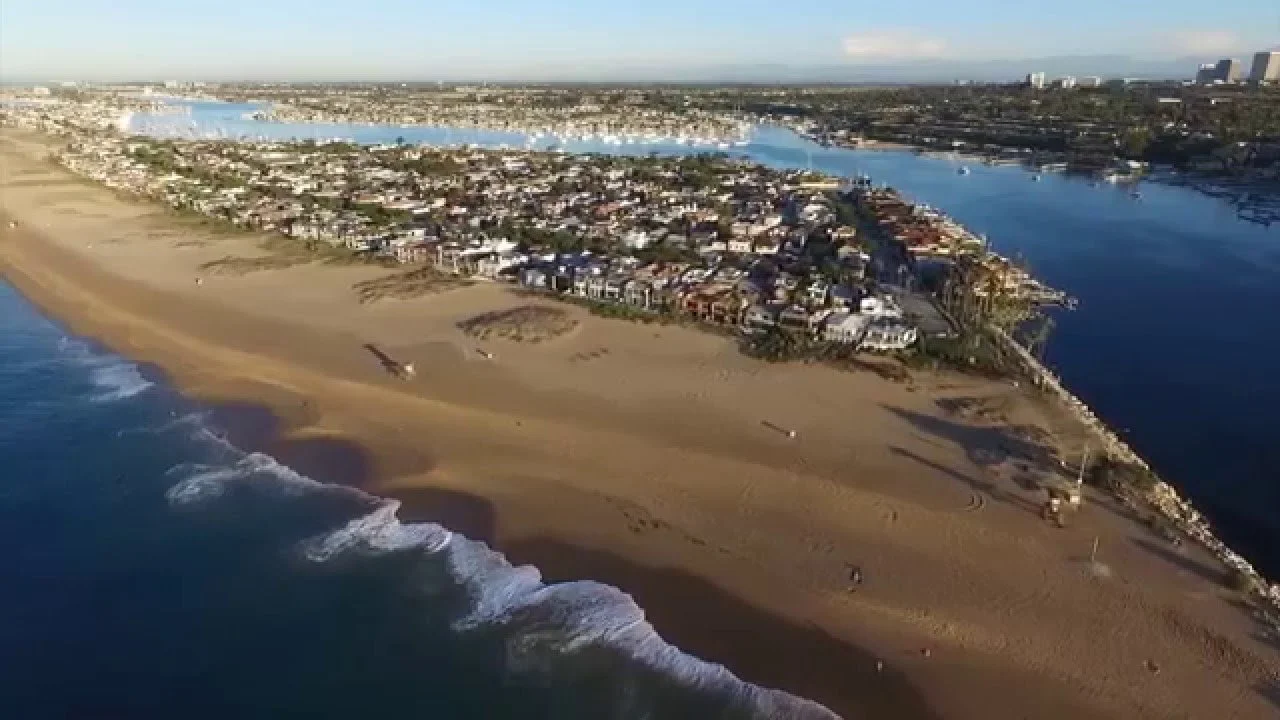 An aerial view of the newport beach peninsula looking right at the world famous wedge next to a residential area and a body of water, with a city skyline in the background.