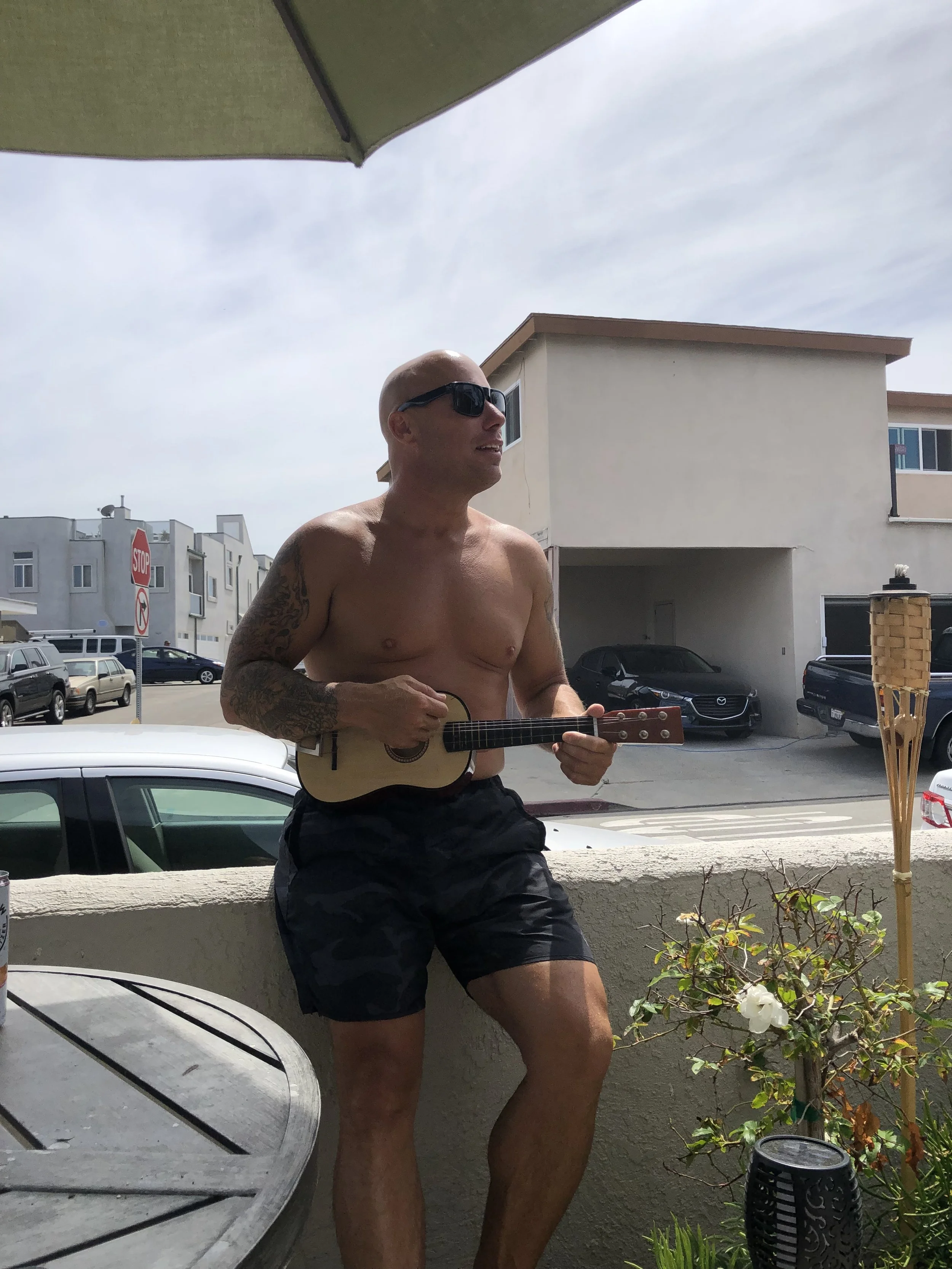 A shirtless man wearing sunglasses and camouflage shorts, holding a small guitar, standing on a balcony with a plant and tiki torch beside him, with buildings and parked cars in the background.