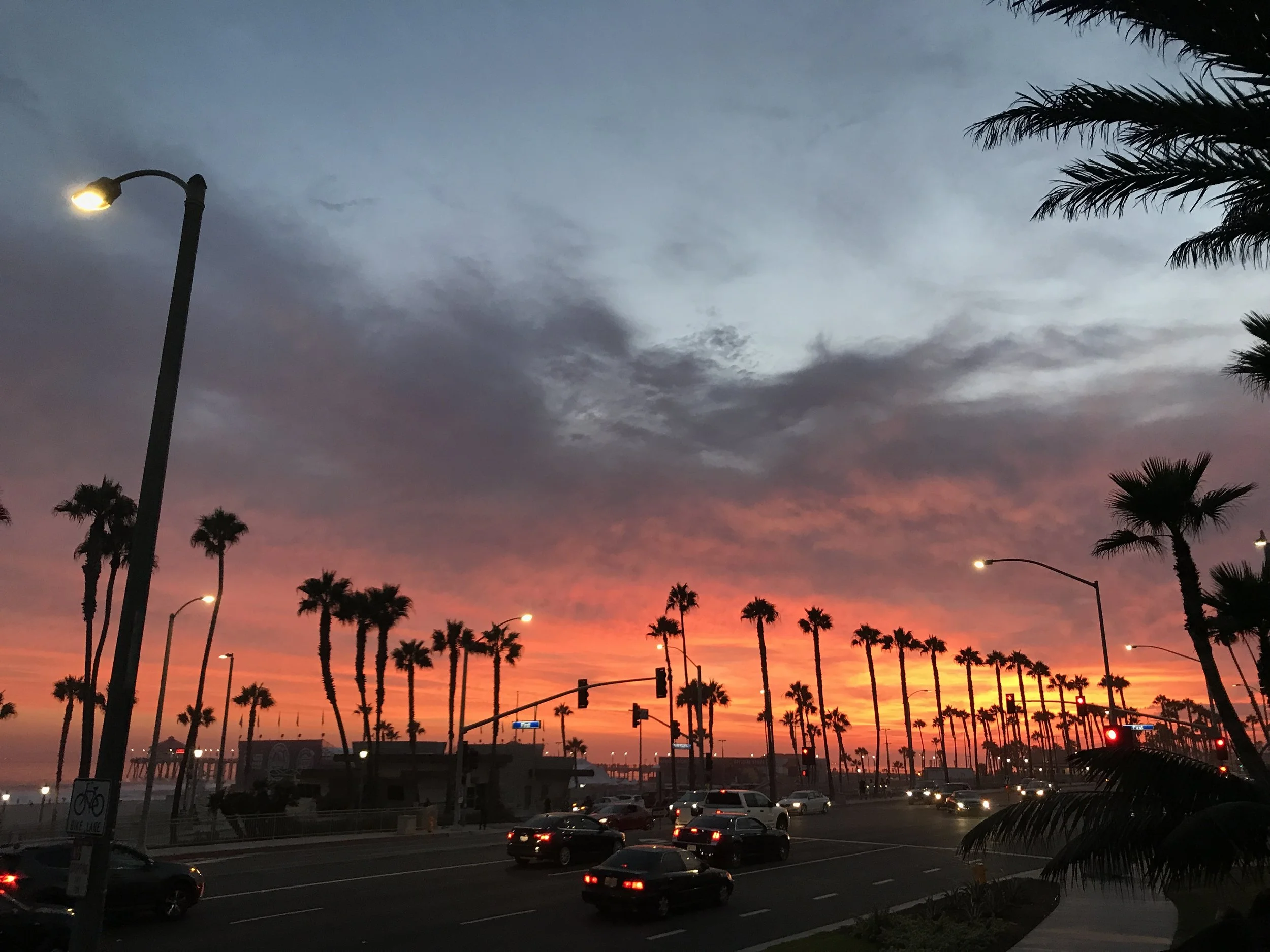 A sunset scene overlooking costa mesa california with a colorful sky, palm trees along the road, streetlights, and traffic on the street.