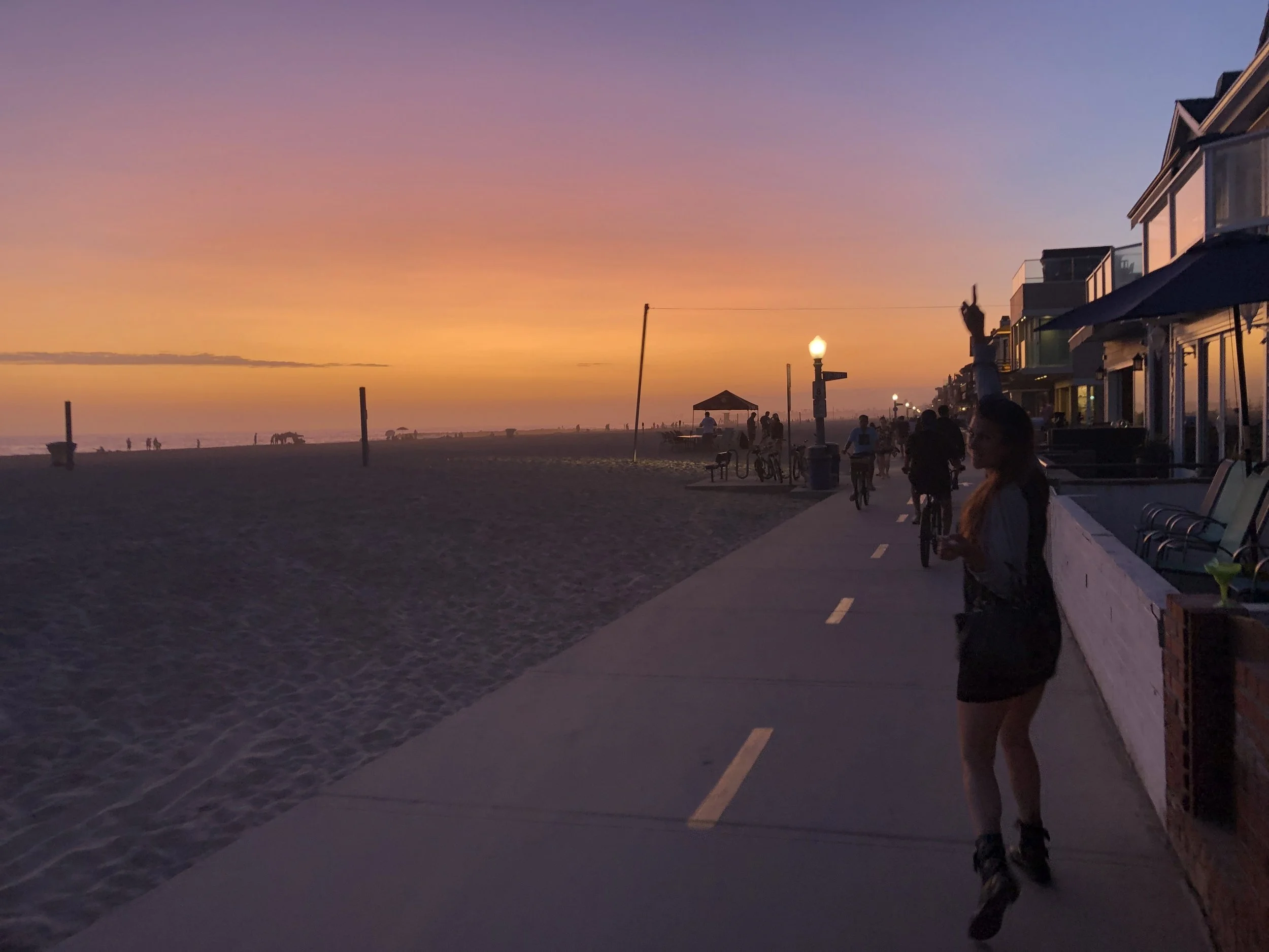 A beachside promenade during sunset with pink and purple sky, people walking, bicycling, and sitting, with beach houses, benches, and streetlamps along the walkway.