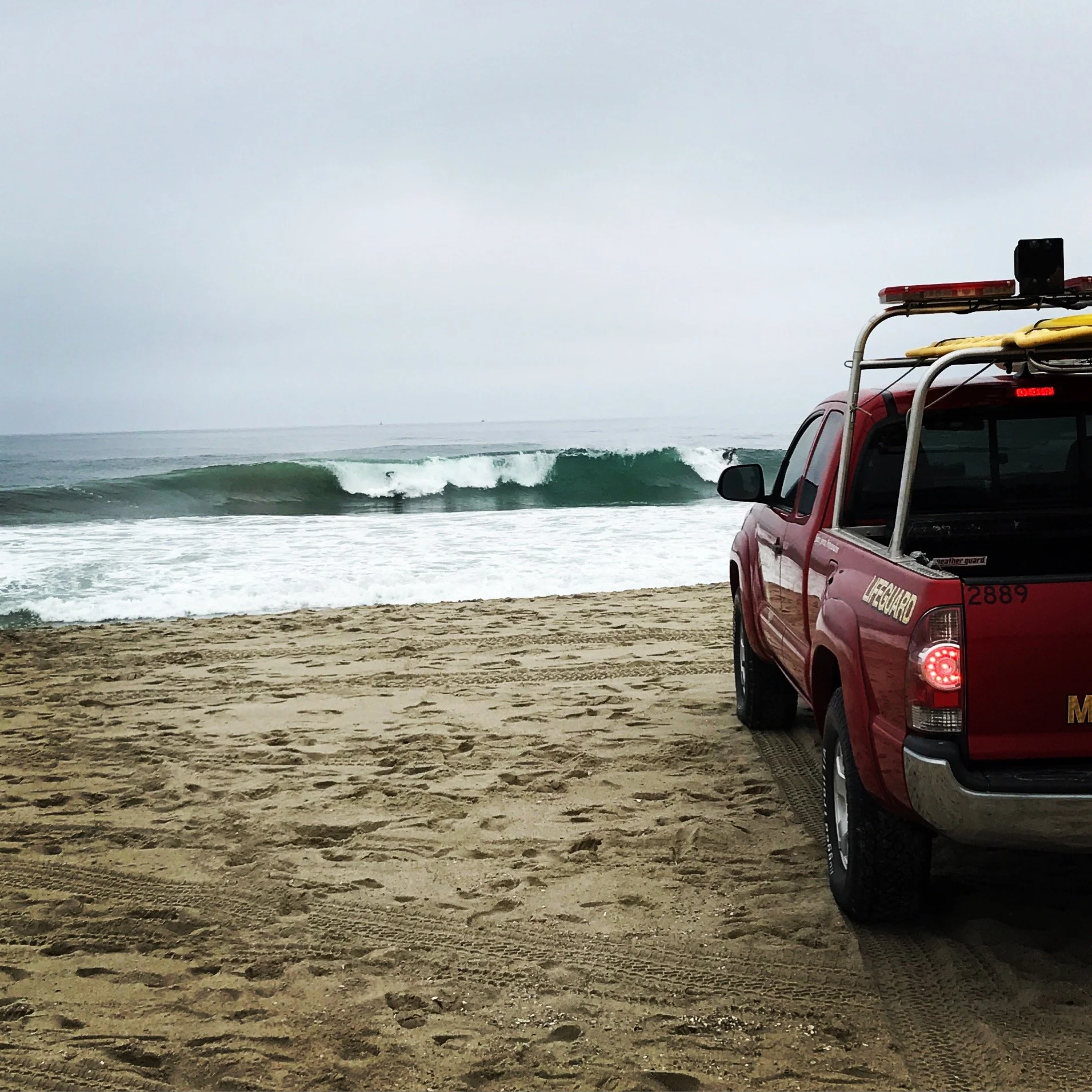 A newport beach lifeguard rescue vehicle parked on a newport beach with a surfboard on its roof, overlooking the ocean with waves and surfer in the background.