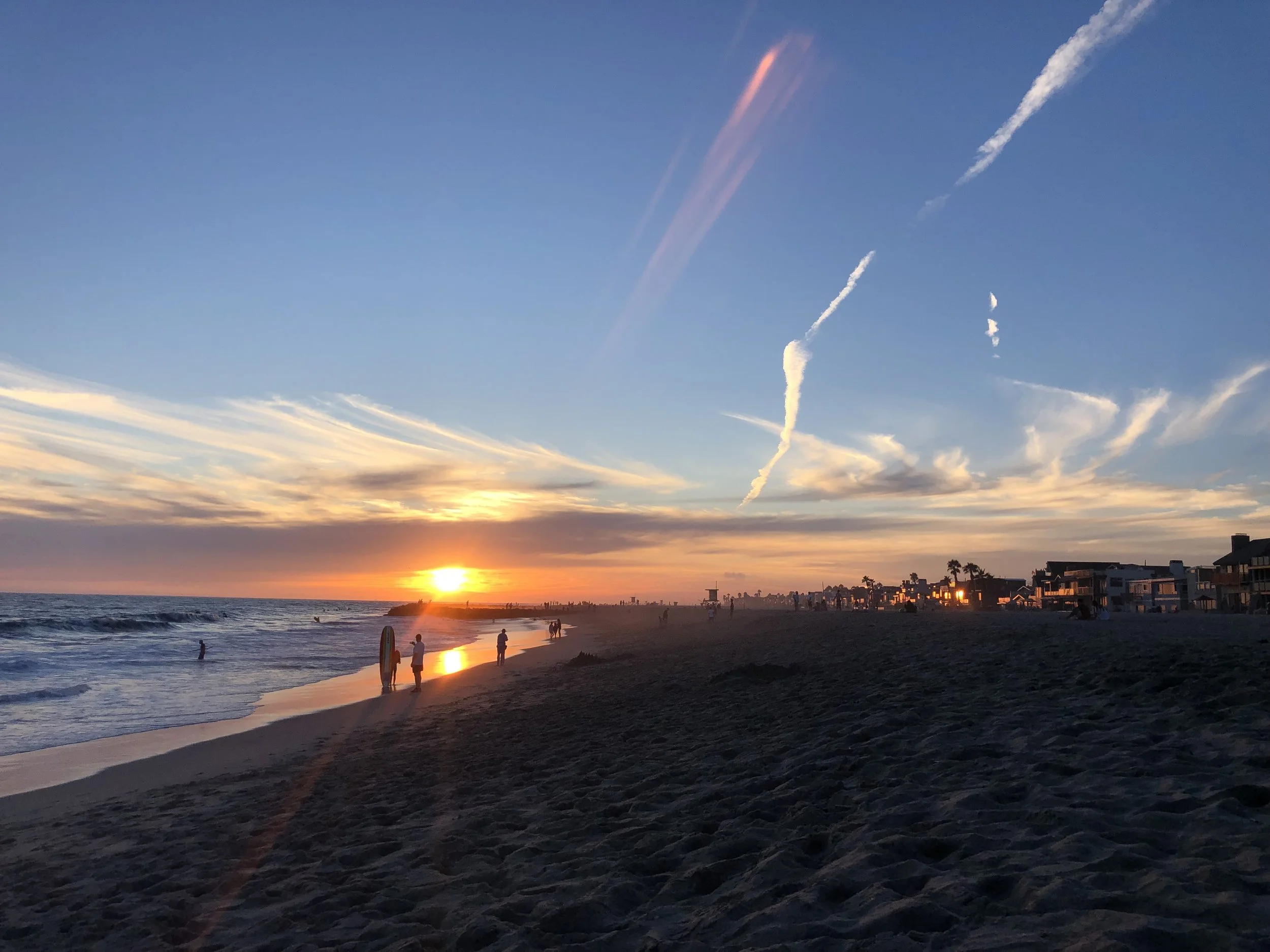 A sunset at the beach with people walking along the shoreline, buildings in the distance, and several contrails in the sky from airplanes.