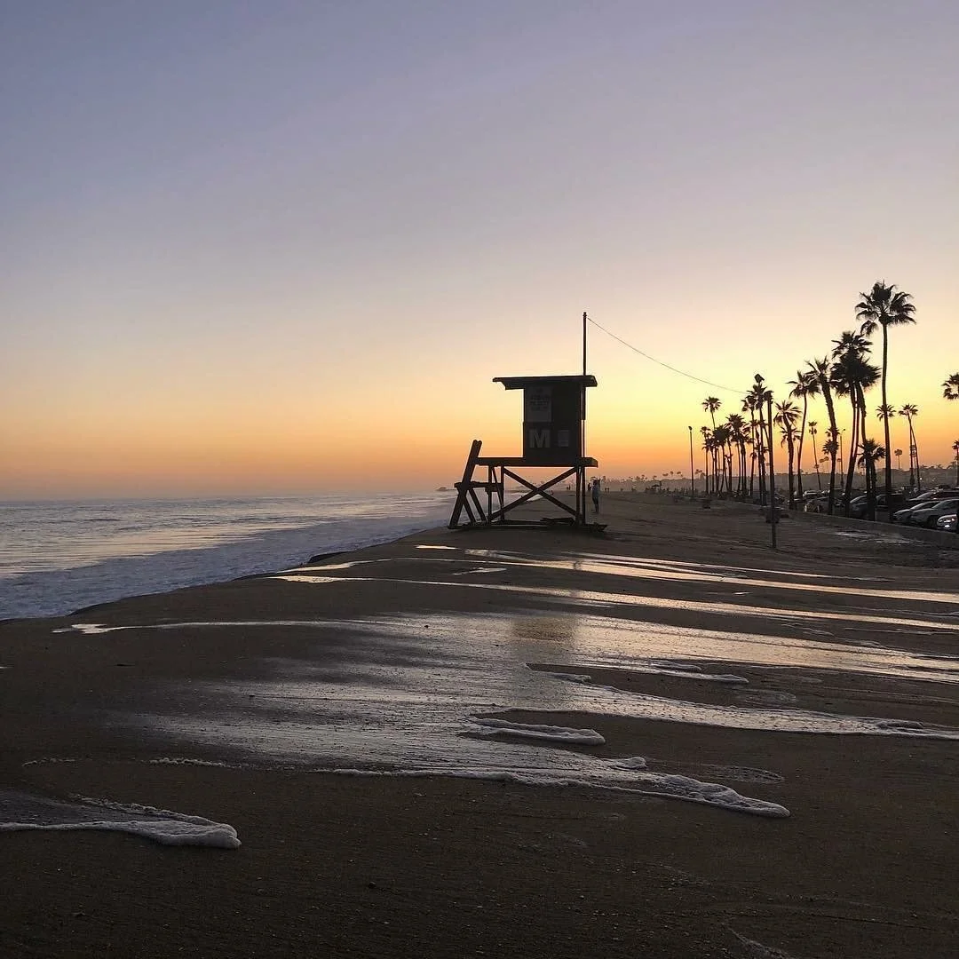 Sunset over a beach with a lifeguard tower, palm trees, and parked cars along the shoreline.