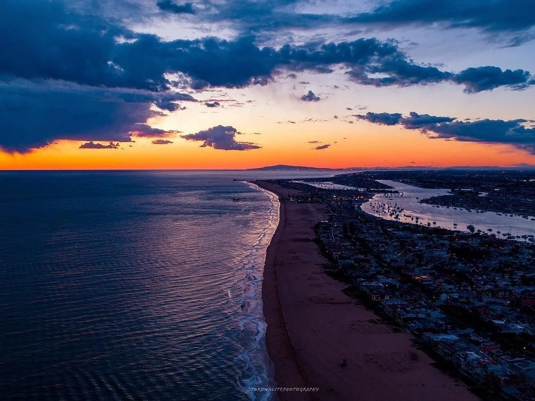 An aerial view of a coastal area at sunset showing a long sandy beach, ocean waves, a city with roads and buildings, and a river flowing into the ocean, with a colorful sky of orange, purple, and blue clouds.
