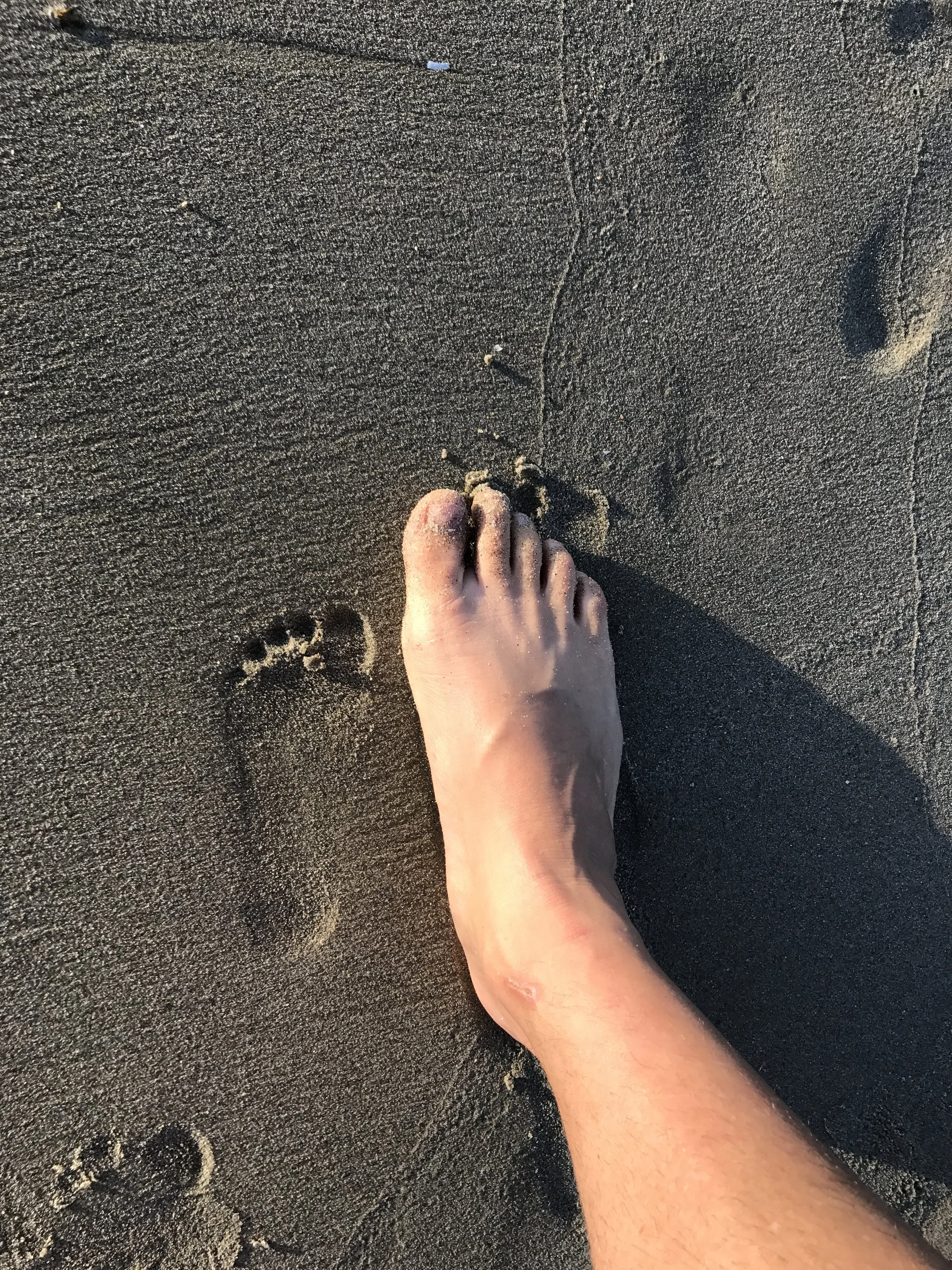 A person's foot and lower leg dug into the sand in newport beach near the river jetties, with footprints and sand patterns around.