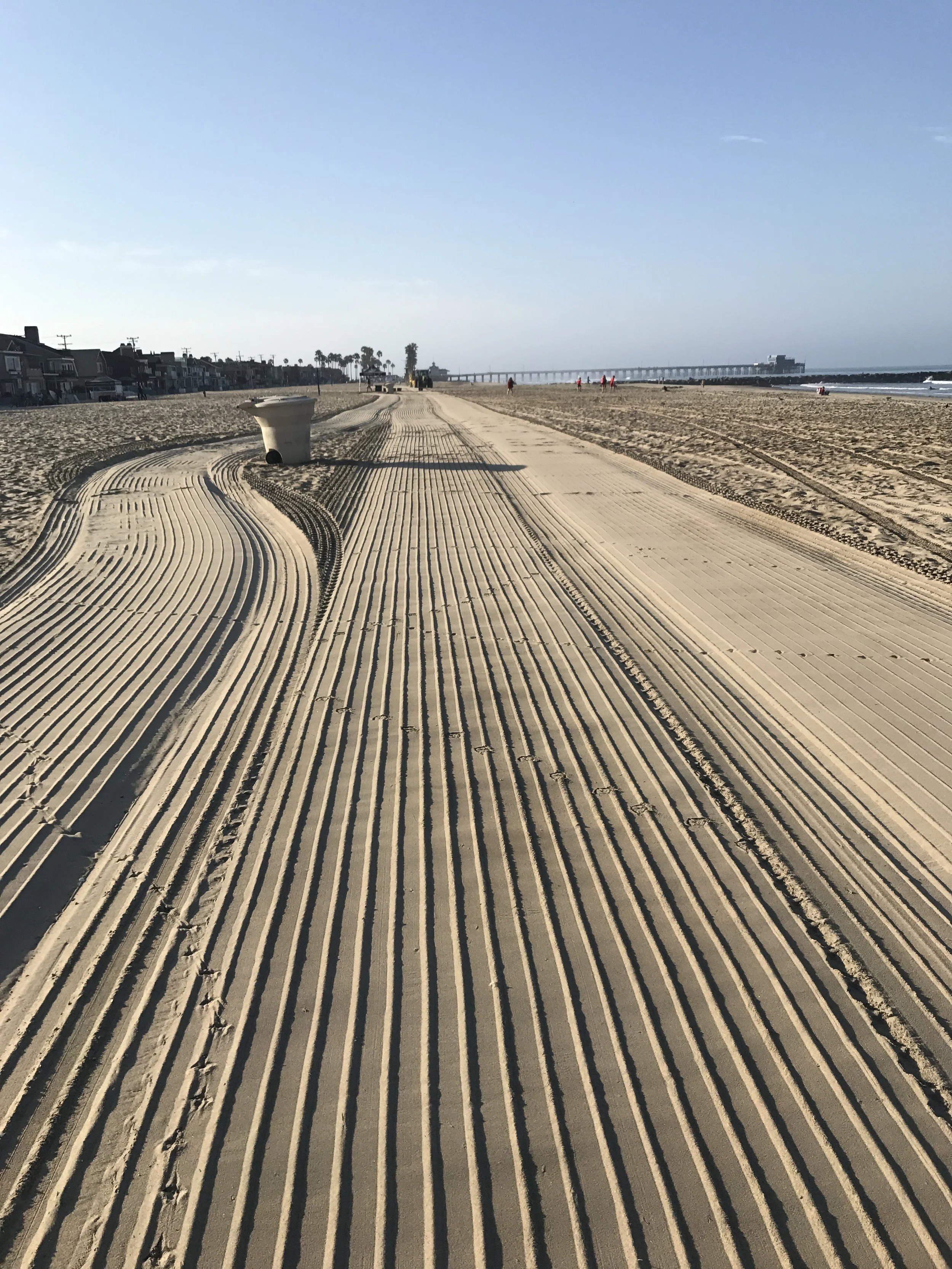 Empty newport beach california with tire track patterns, a distant pier, and a few people walking, under a clear blue sky.