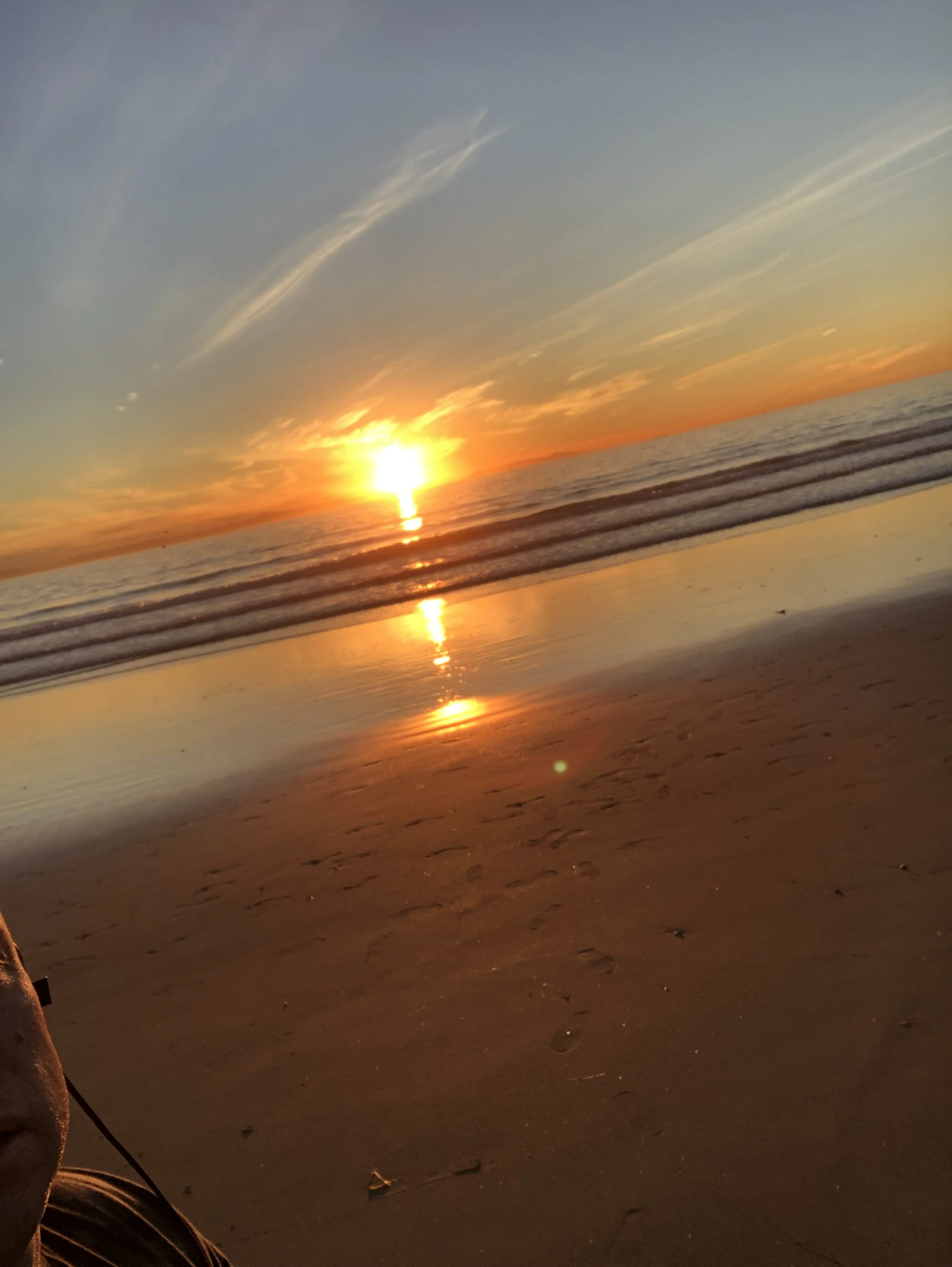Sunset over the ocean at a beach, with the reflection of the sun on the water and a clear sky with some clouds.