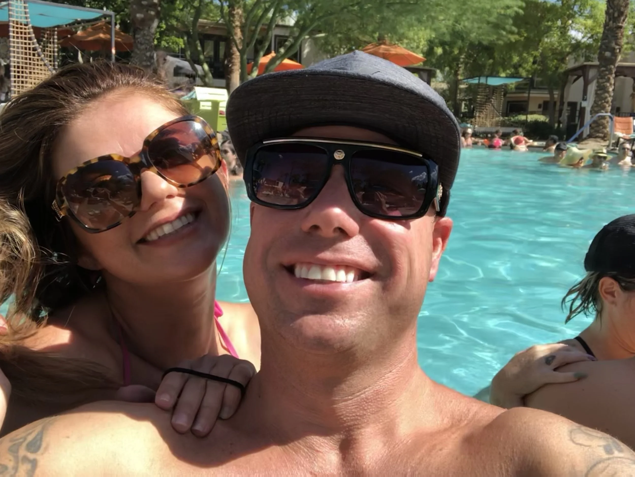 Smiling woman with curly hair wearing large sunglasses, standing close to a man with a big smile, dark hat, and sunglasses, at a poolside with trees, umbrellas, and people in the background.