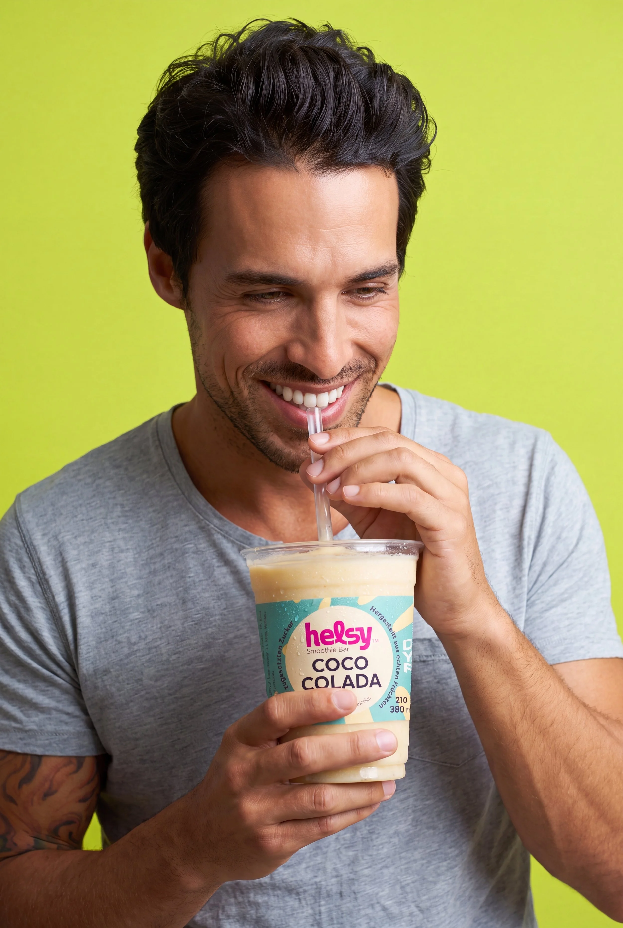 Mann mit Tattoos trinkt einen Kokos-Coloda-Smoothie aus einem Becher mit rotem Strohhalm vor grünem Hintergrund.
