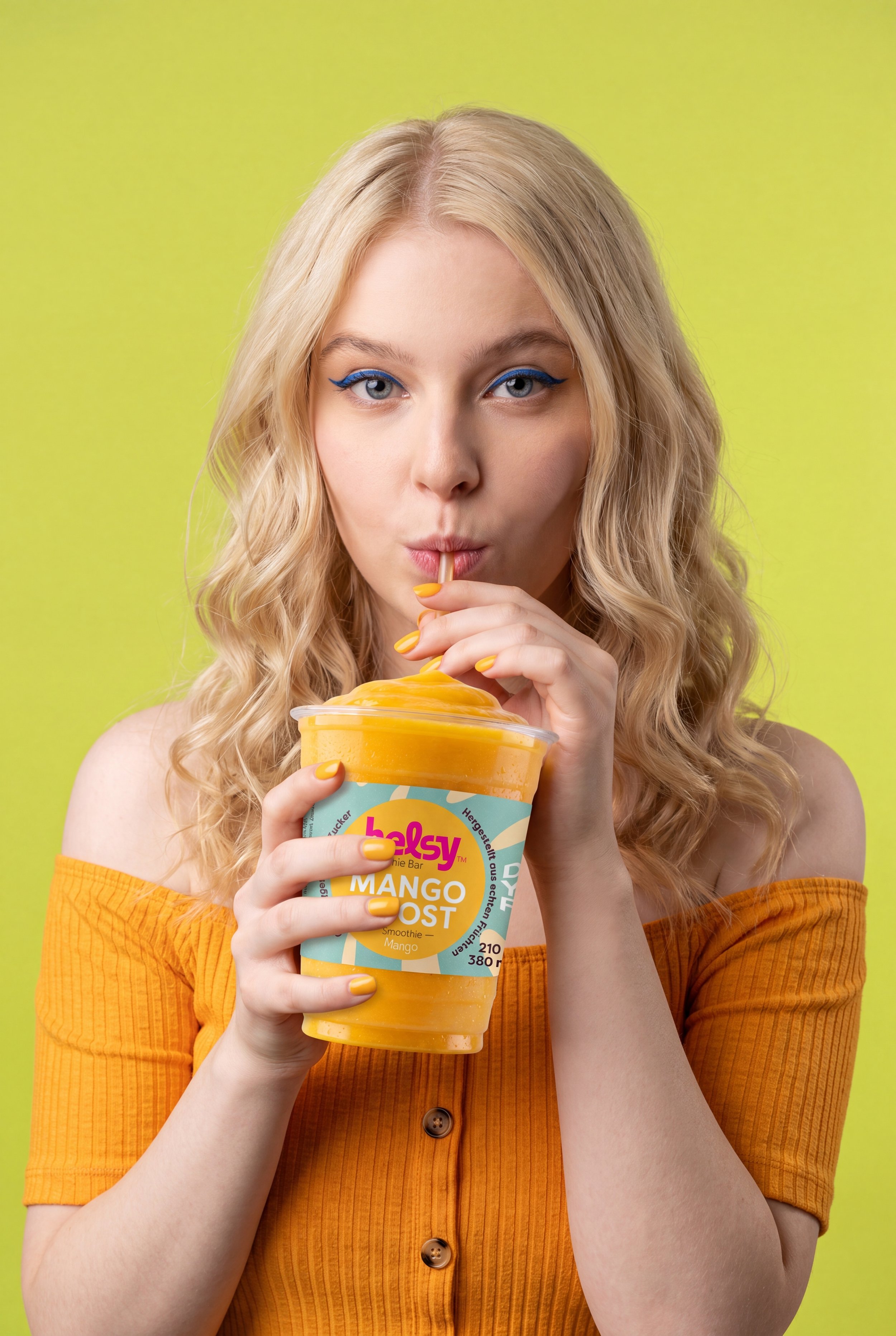 Junge Frau mit blonden, lockigen Haaren und blauen Augen, die Mango-Smoothie trinkt, trägt ein orangefarbenes Oberteil mit Knöpfen, vor hellgrünem Hintergrund.