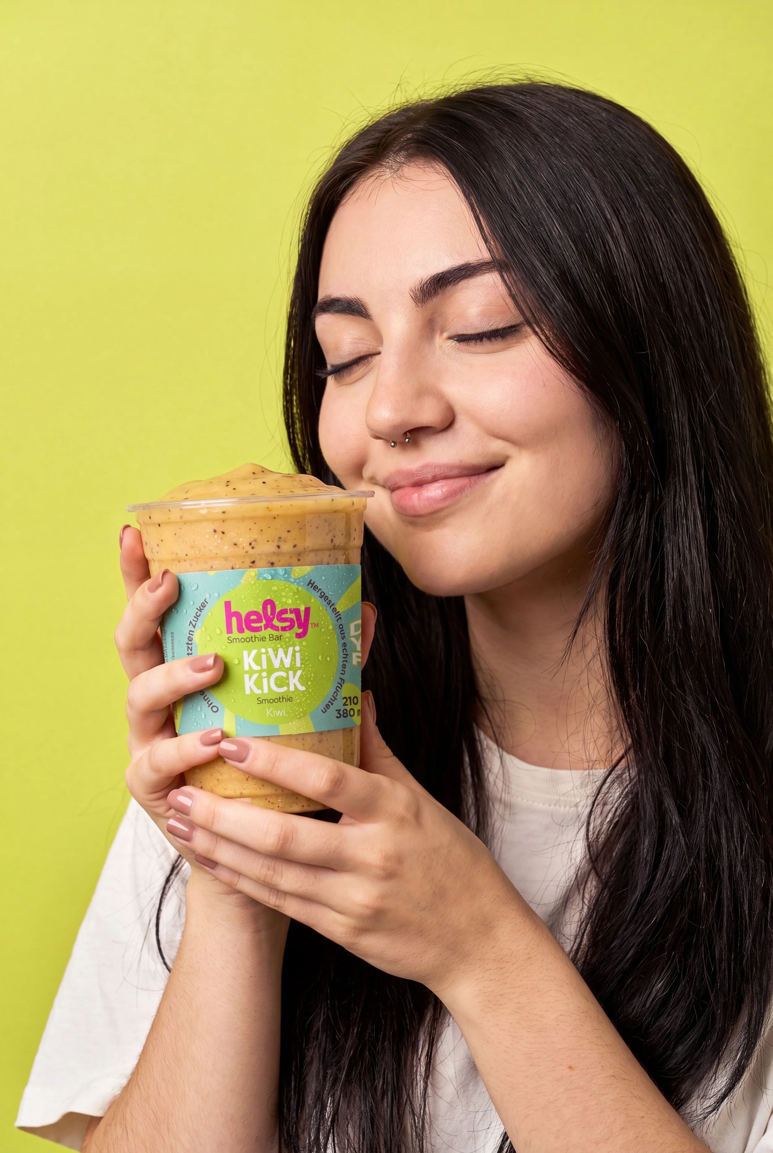 Junge Frau mit glatter, dunkler Haare hält eine Kiwi-Kick Smoothie in ihrer Hand, die ihr Gesicht fast berührt, vor hellgrüner Wand, trägt weißes T-Shirt.