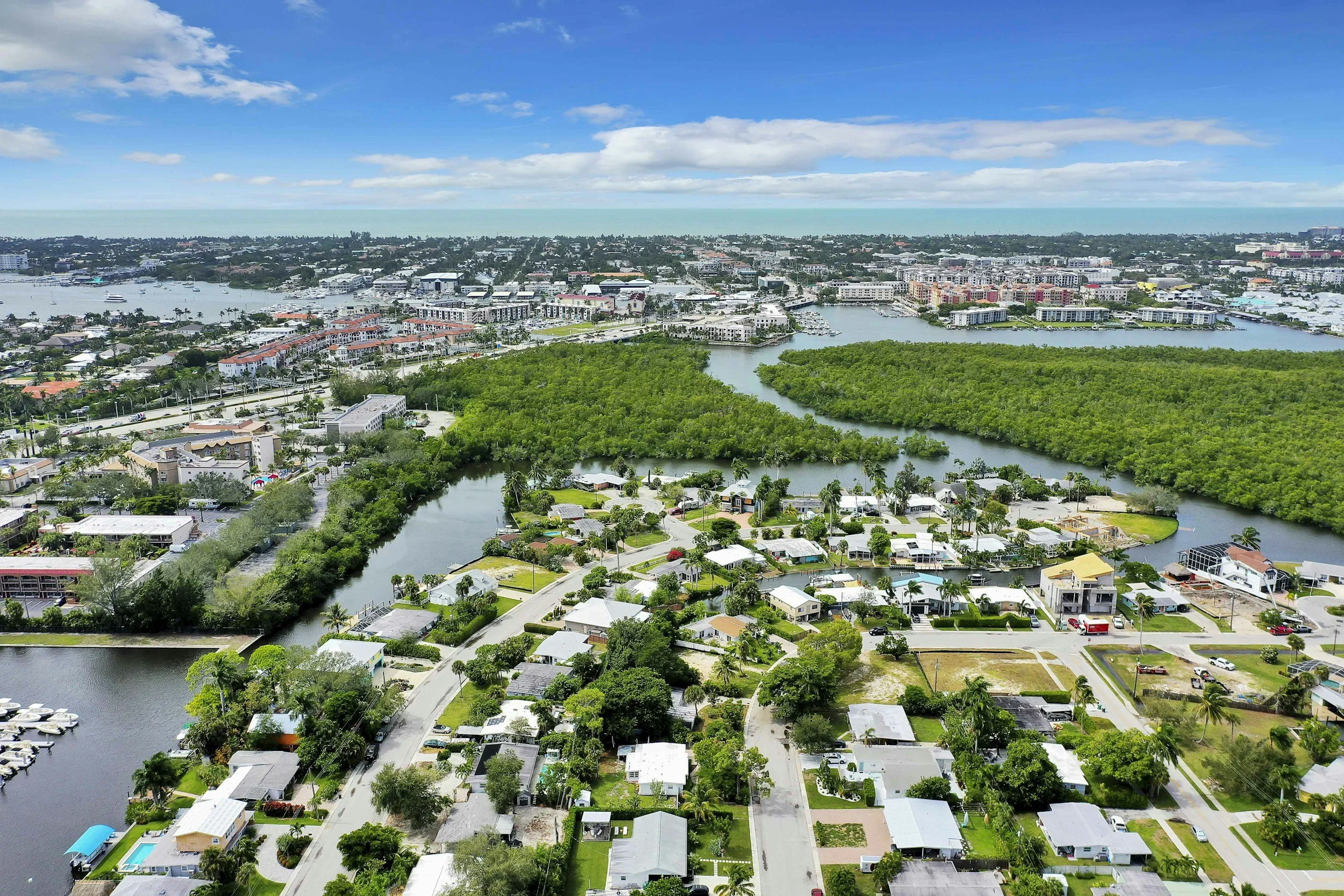 Aerial view of a coastal city with waterway, boats, residential neighborhoods, and commercial buildings under a partly cloudy sky.