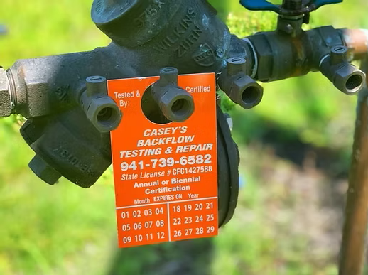 A black outdoor water valve with a red casing tag attached. The tag reads "Casey's Backflow Testing & Repair" and provides a phone number, certification details, and an expiration date, with numbered months listed for the year.