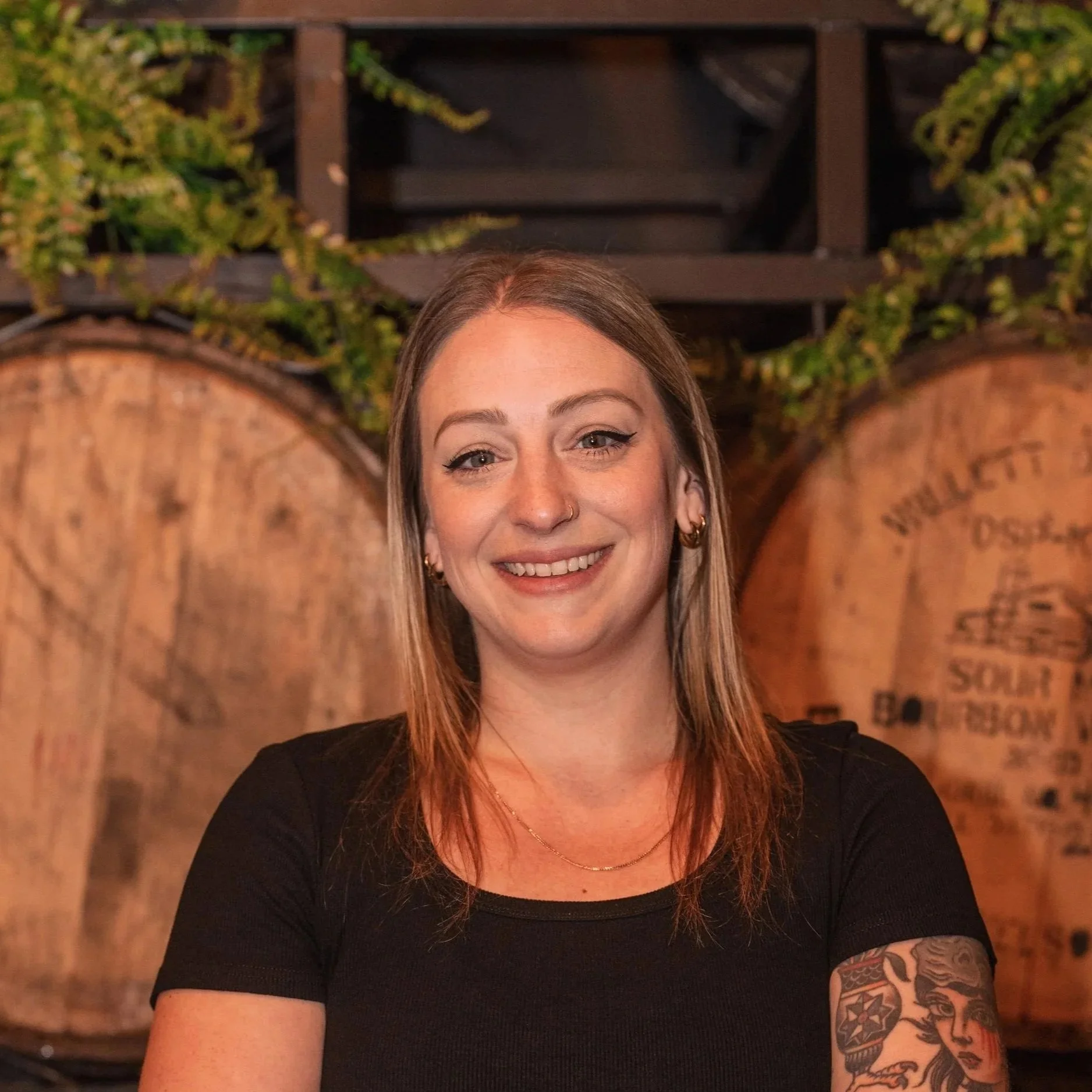 A woman with light skin and long, light brown hair smiling. She has a nose ring, wears earrings, a necklace, and a black shirt. She has a tattoo sleeve on her right arm. The background features a wooden barrel and greenery.