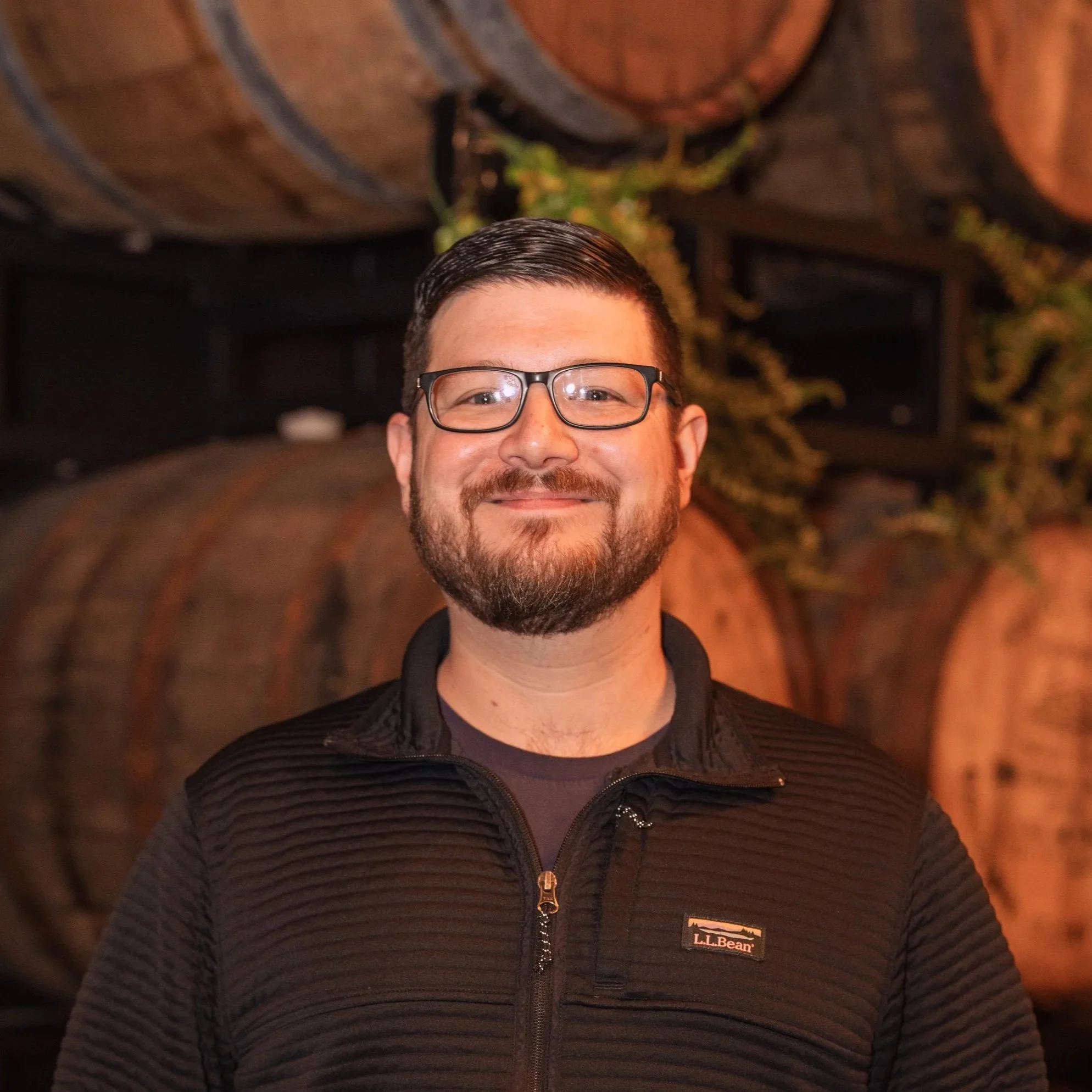 A man with glasses and a beard smiling, wearing a black L.L. Bean jacket, standing outdoors at night with trees in the background.
