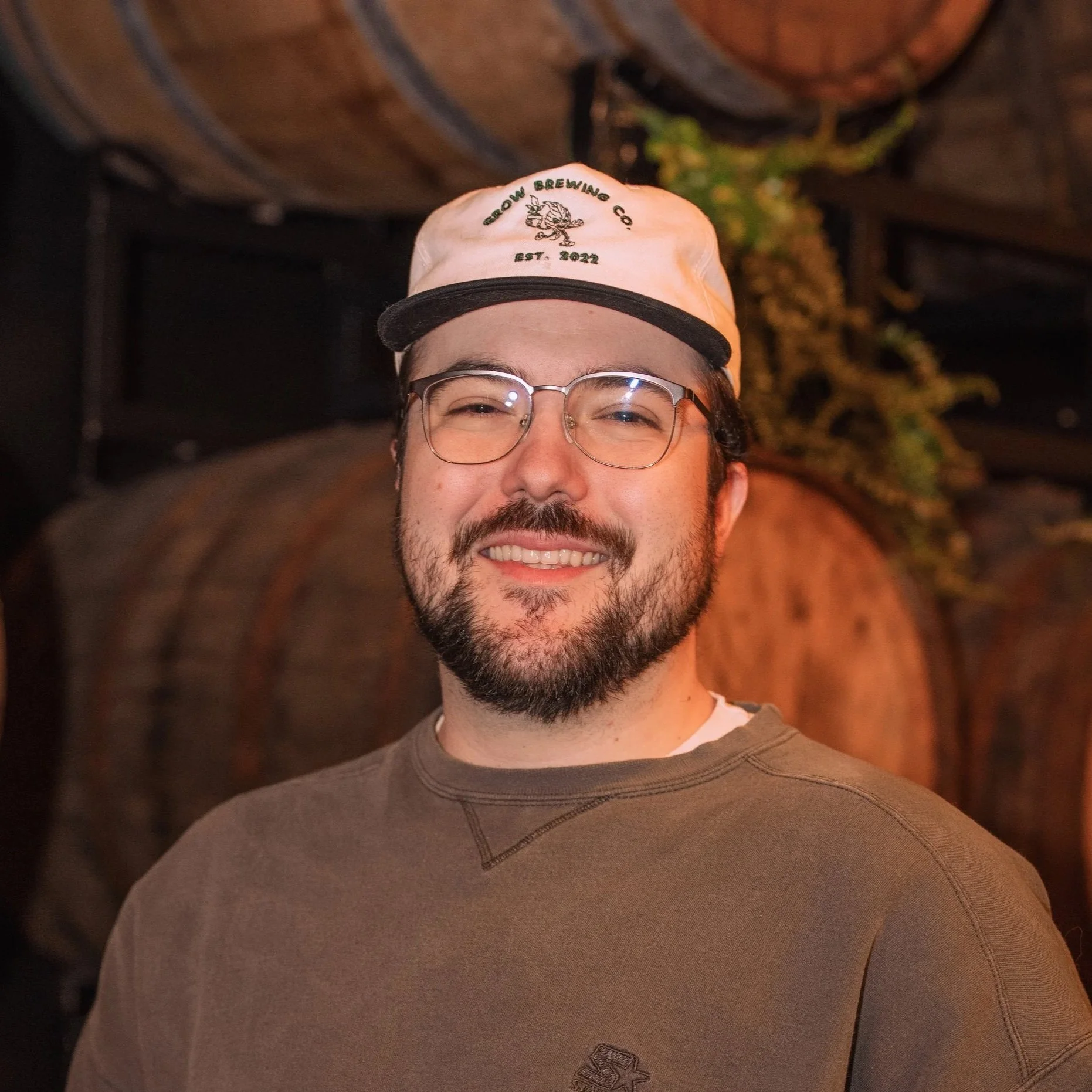 A smiling man with glasses wearing a beige cap with green embroidery reading 'Grow Brewing Co. Est. 2022'. He is wearing a brown shirt and has a beard. The background is dark with some blurred plants and a wooden structure.