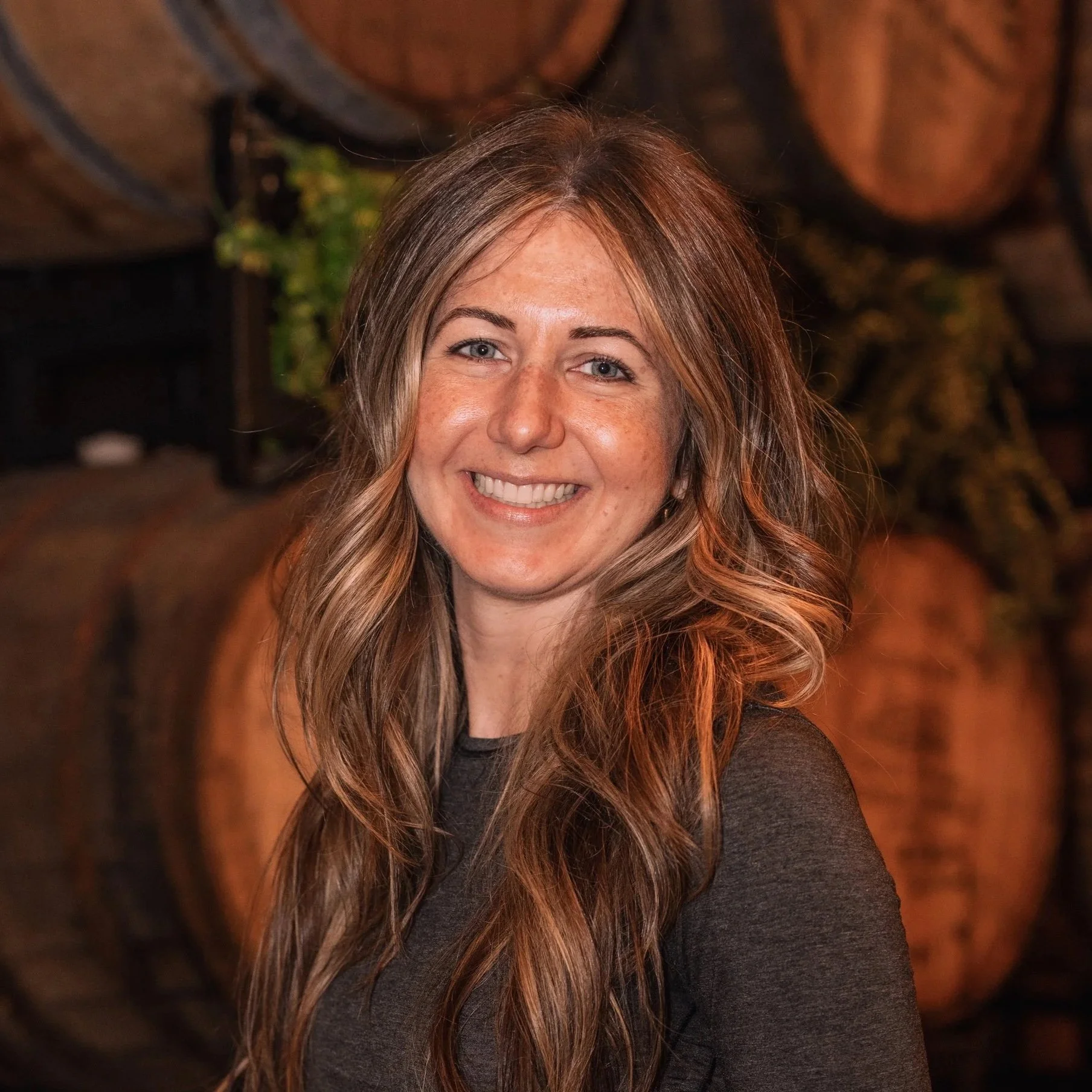A woman with long, wavy, light brown hair and blue eyes smiling, wearing a dark top, with a background of warm-toned blurred decorations.