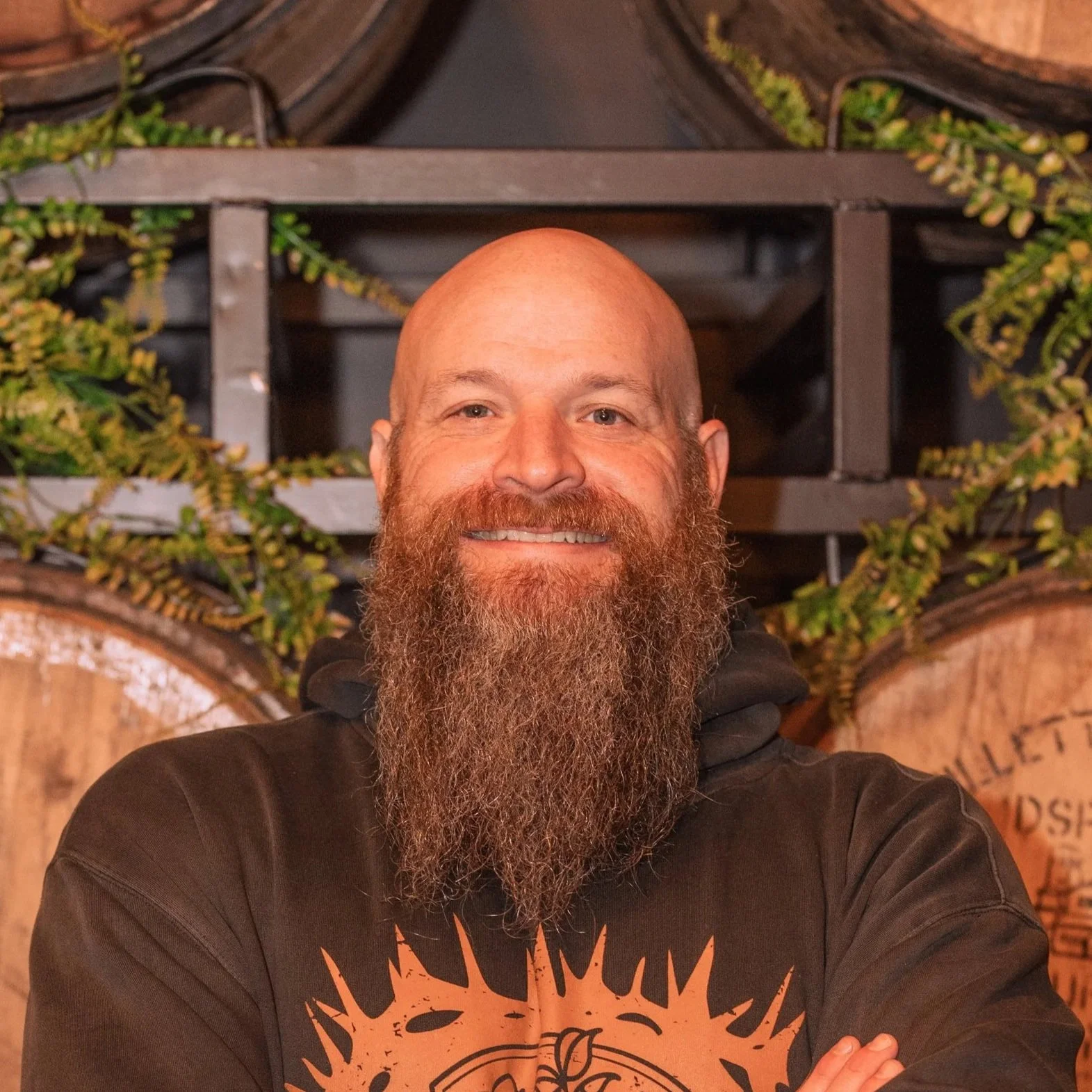 A man with a bald head and long beard smiling and taking a selfie in front of a wall decorated with horizontal wooden barrels and green plants.