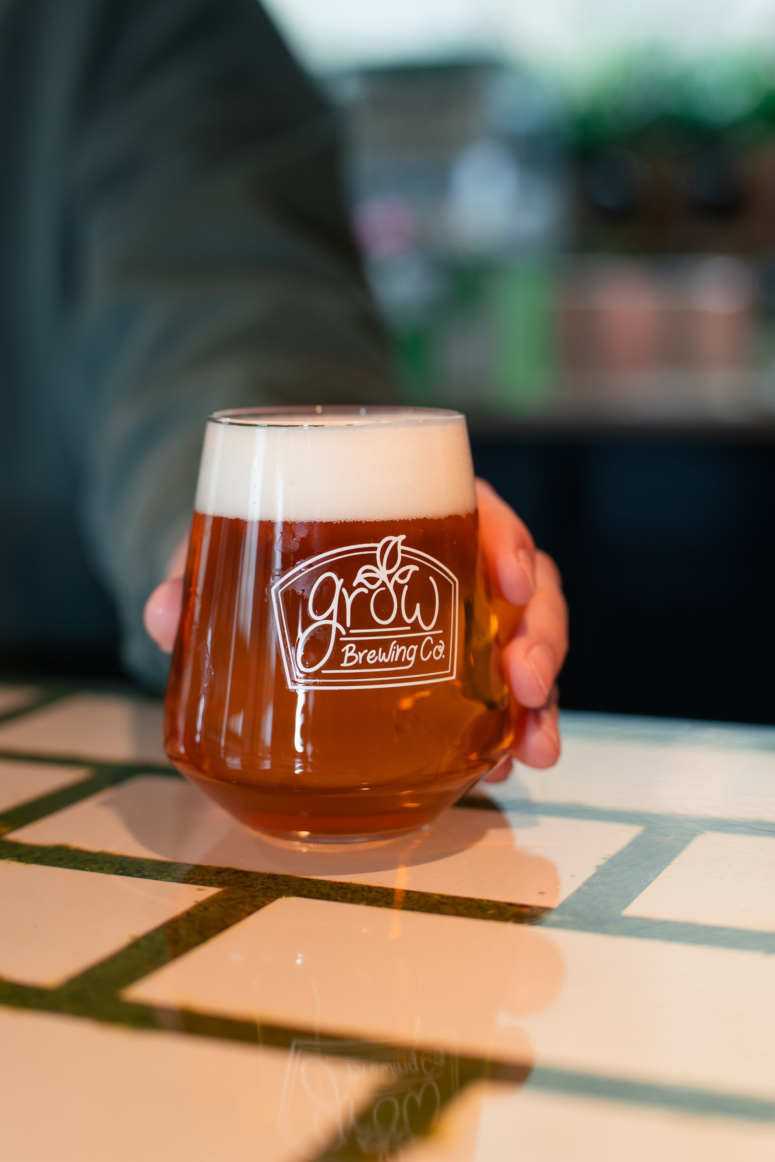 A person holding a glass of beer with the logo 'Grow Brewing Co' on it, sitting on a tiled surface with the bottle's reflection visible.
