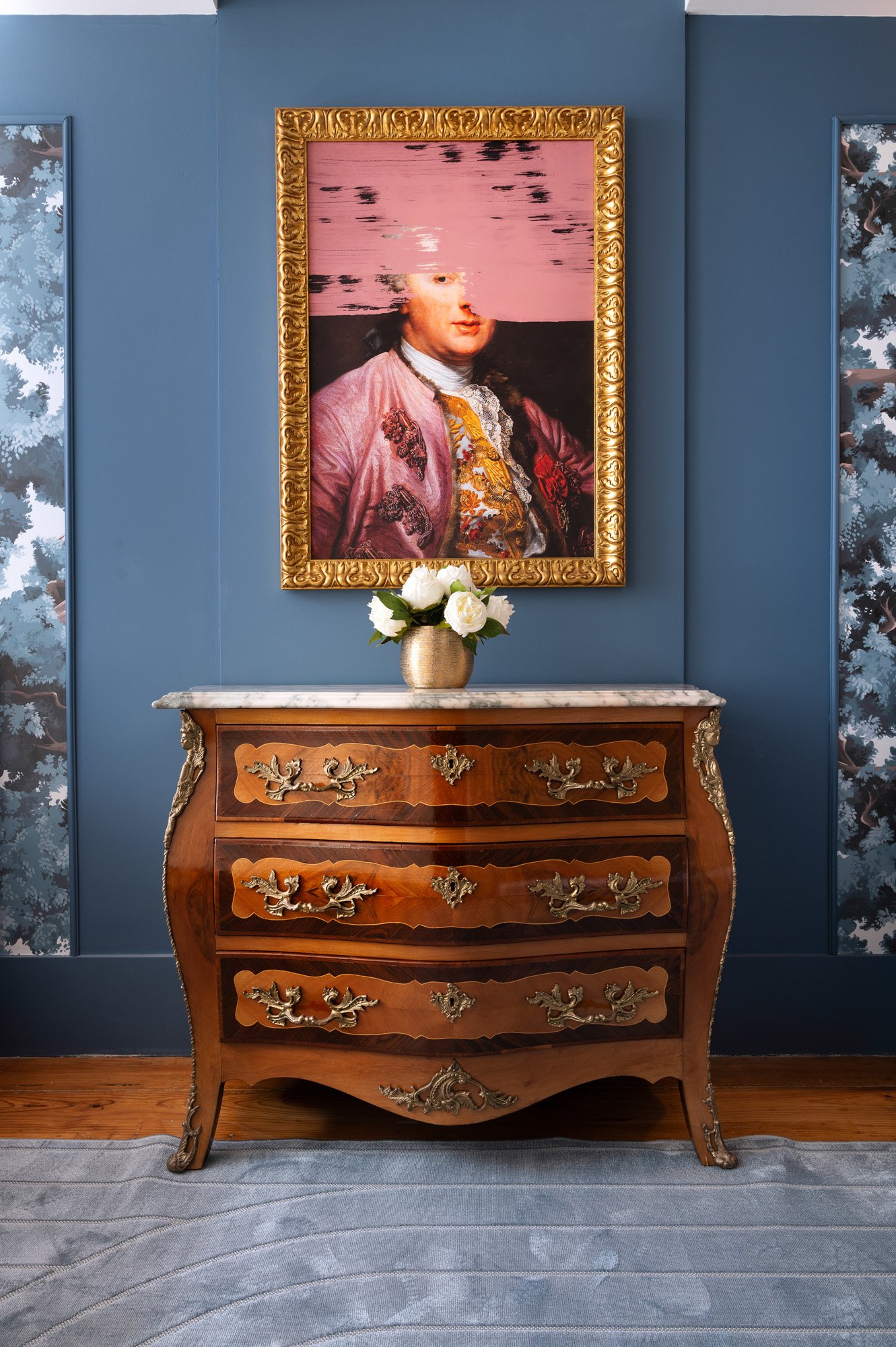 A vintage wooden dresser with ornate gold and silver decorative details, placed in front of a blue wall with framed wallpaper on either side. A gold vase with white roses sits on top of the dresser. Above it, a framed portrait painting of a woman with a sky and water scene superimposed over her face.
