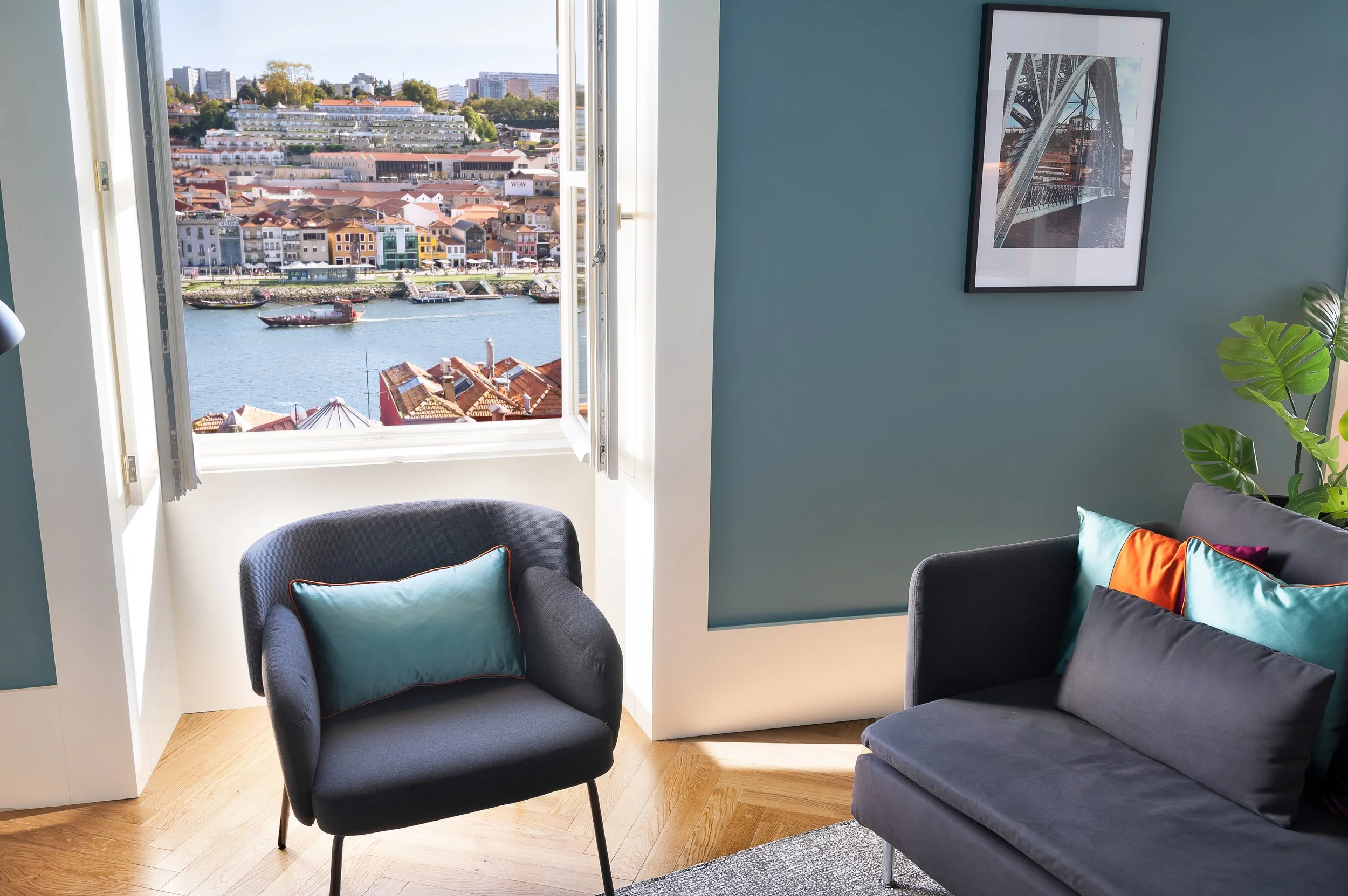 Living room with large window overlooking a river and cityscape, blue accent wall with framed cityscape photo, dark couch with colorful pillows, black armchair with teal pillow, and green potted plant.
