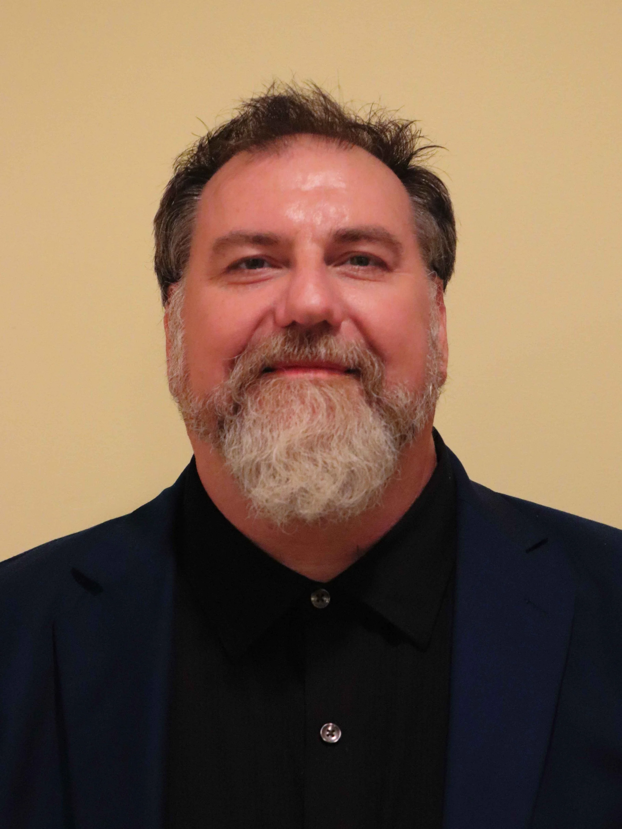 Close-up of a middle-aged man with a beard and mustache smiling, wearing a dark suit jacket and shirt, against a plain beige background.