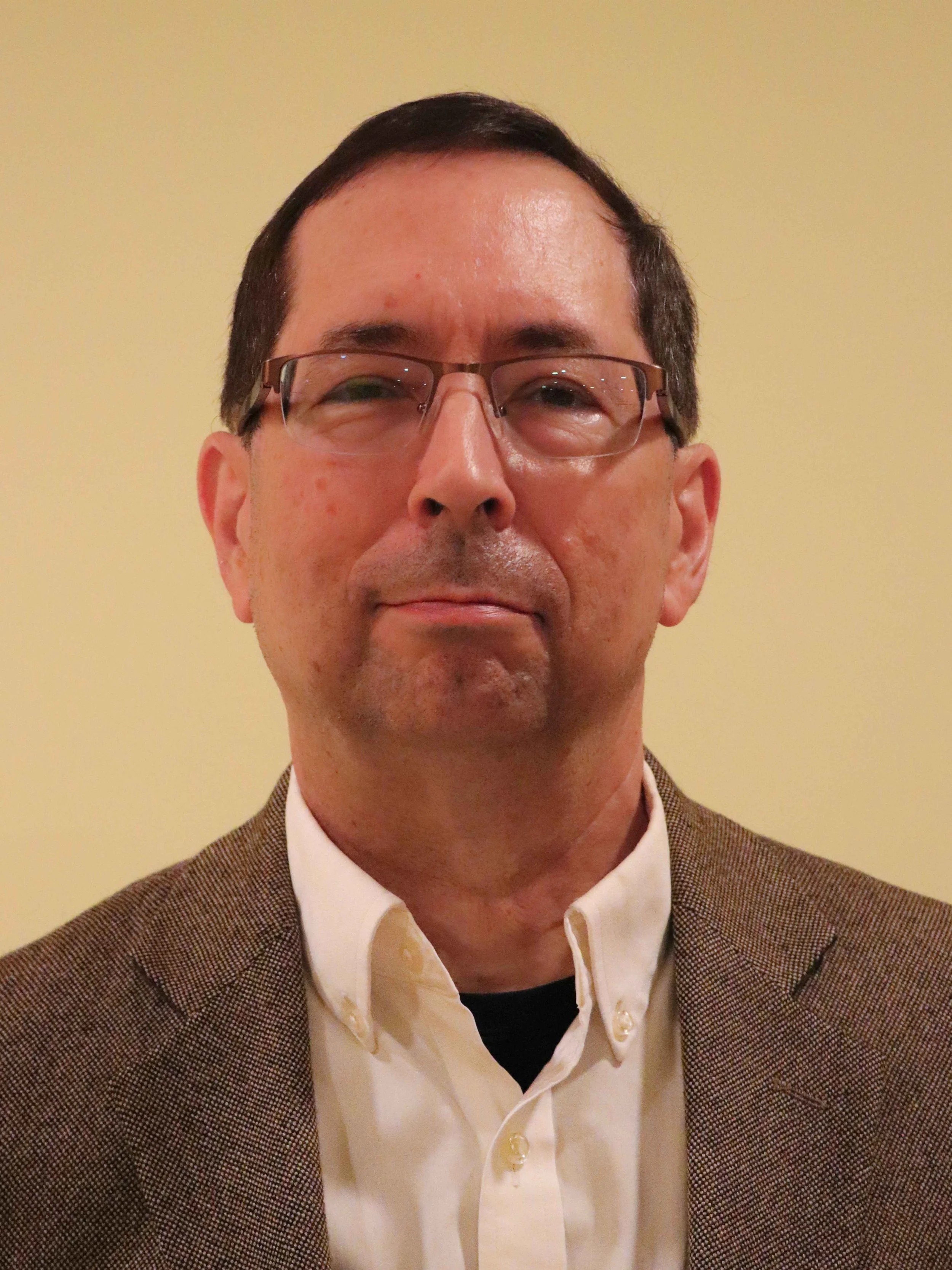 Close-up of a smiling man with glasses, wearing a brown checked blazer and a white shirt, against a plain yellow background.