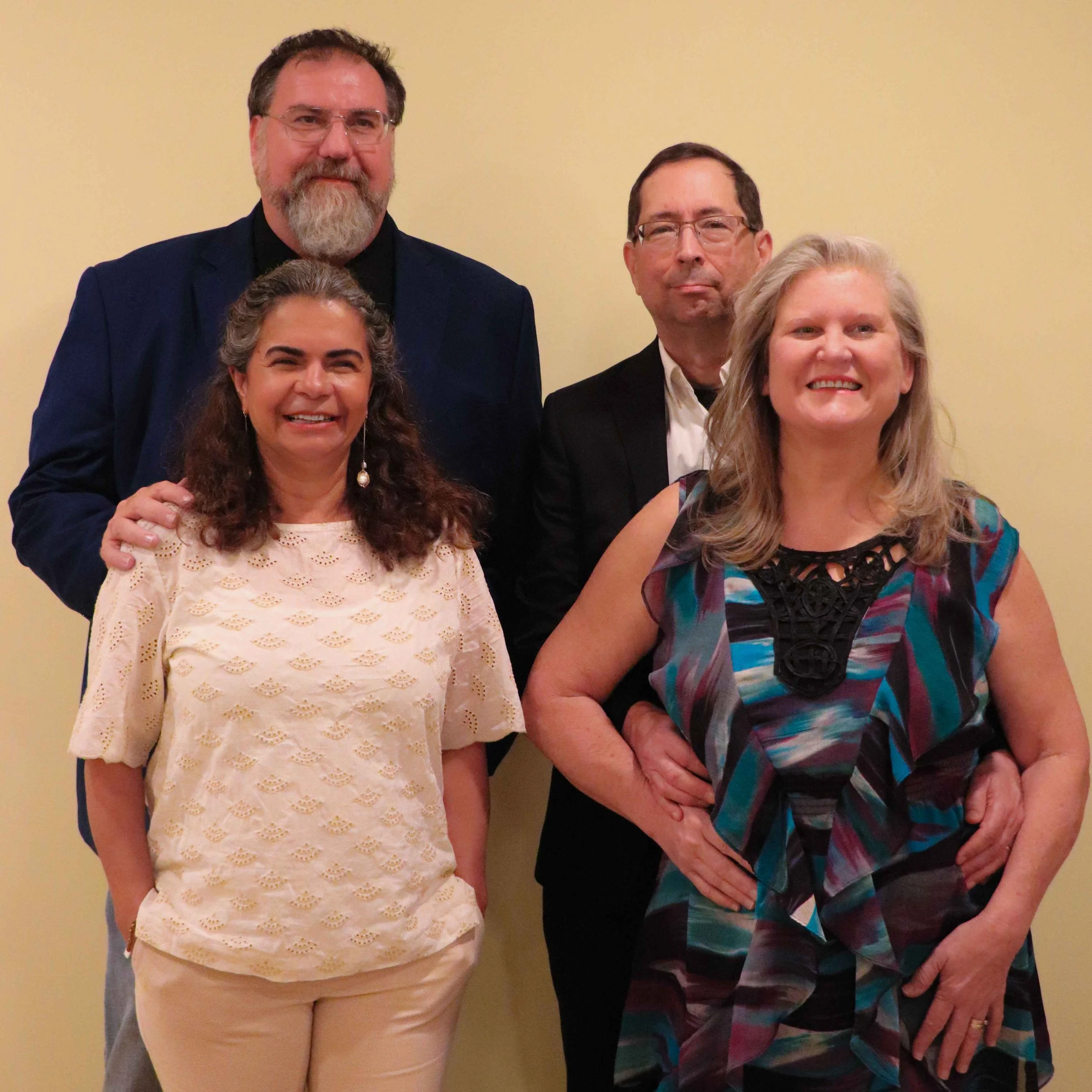 A group of four people, two men and two women, posing against a beige wall, dressed in formal attire, smiling for the photo.