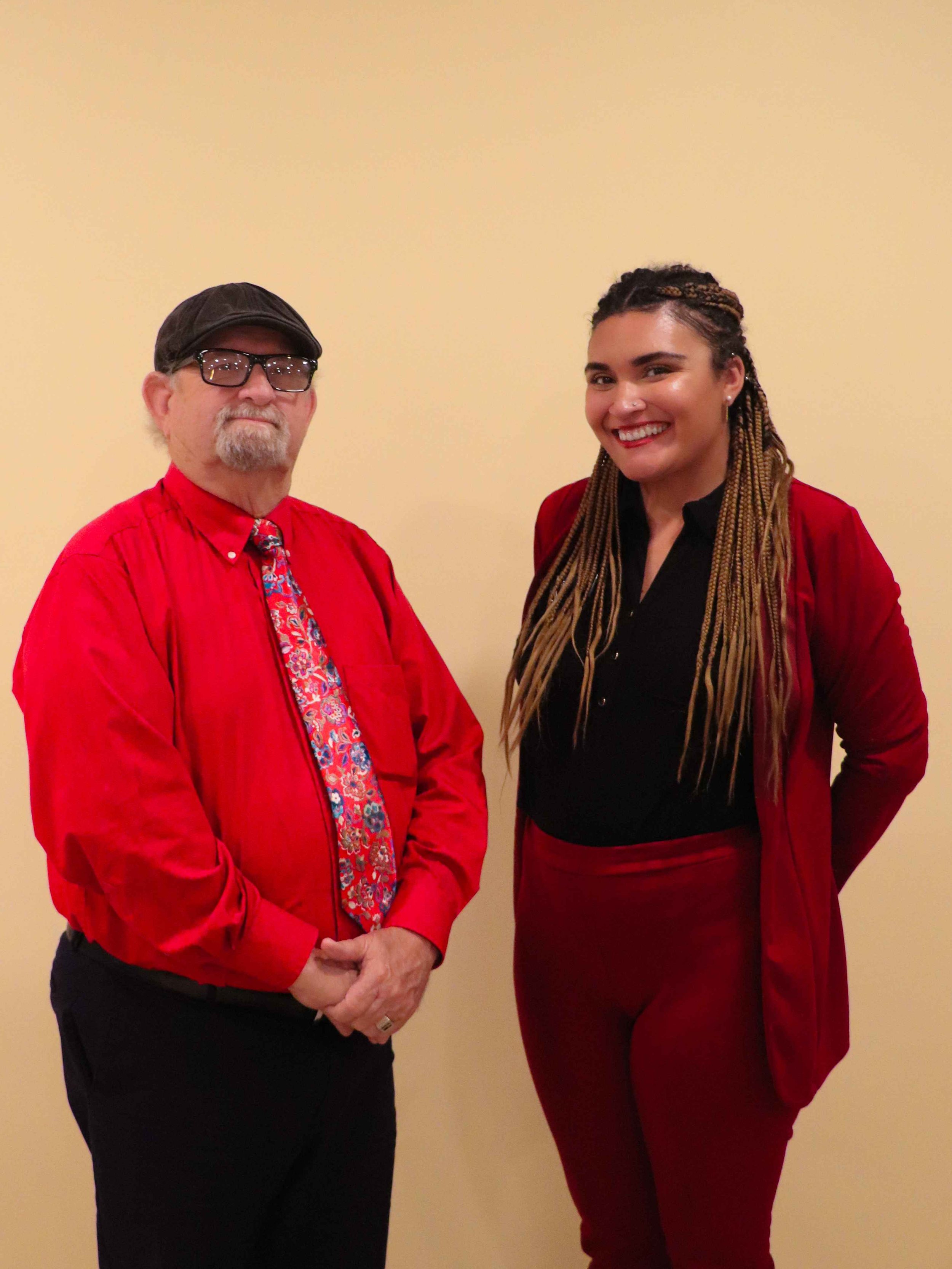 A man and a woman standing against a plain yellow wall, both dressed in red and smiling, with the man wearing glasses and a cap, and the woman with long braided hair.