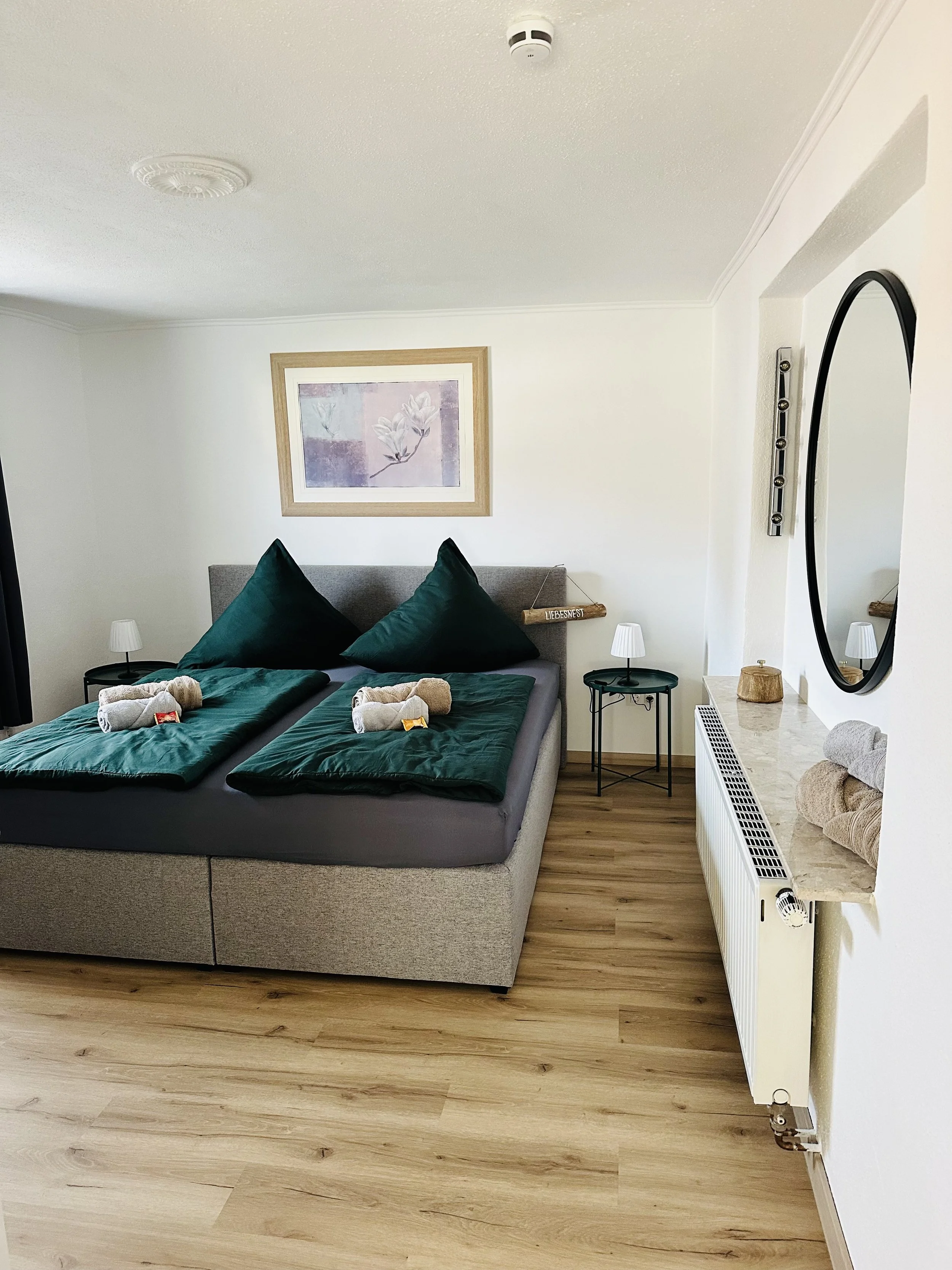 Ein Schlafzimmer mit einem großen Doppelbett, das mit dunkelgrünen Bettwäsche und Kissen bedeckt ist. Auf dem Bett liegen eingerollte Handtücher. Über dem Bett hängt ein Rahmen mit einem Blumenbild. Es gibt zwei kleine Nachttische mit Lampen, ein Wandbild und einen Heizkörper mit einem Spiegel darüber.