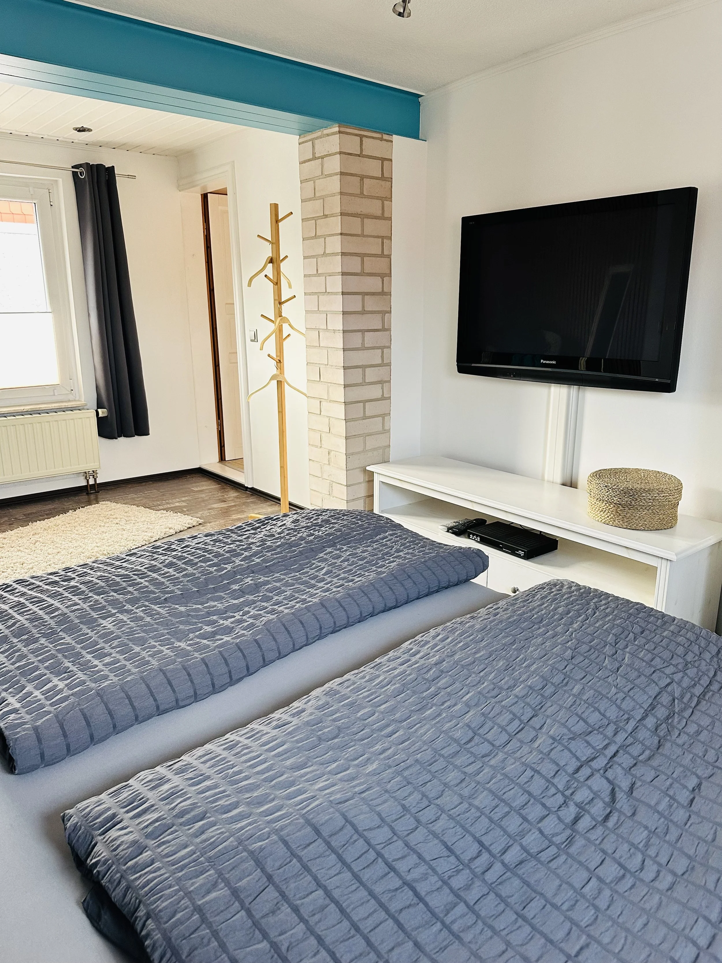 Zwei Betten mit blauen Bettdecken in einem Schlafzimmer, ein Fernseher an der Wand, ein Holzkleiderständer und ein Fenster mit grauer Vorhang. Es gibt auch einen weißen Wandschrank, eine Heizung und einen kleinen Teppich.