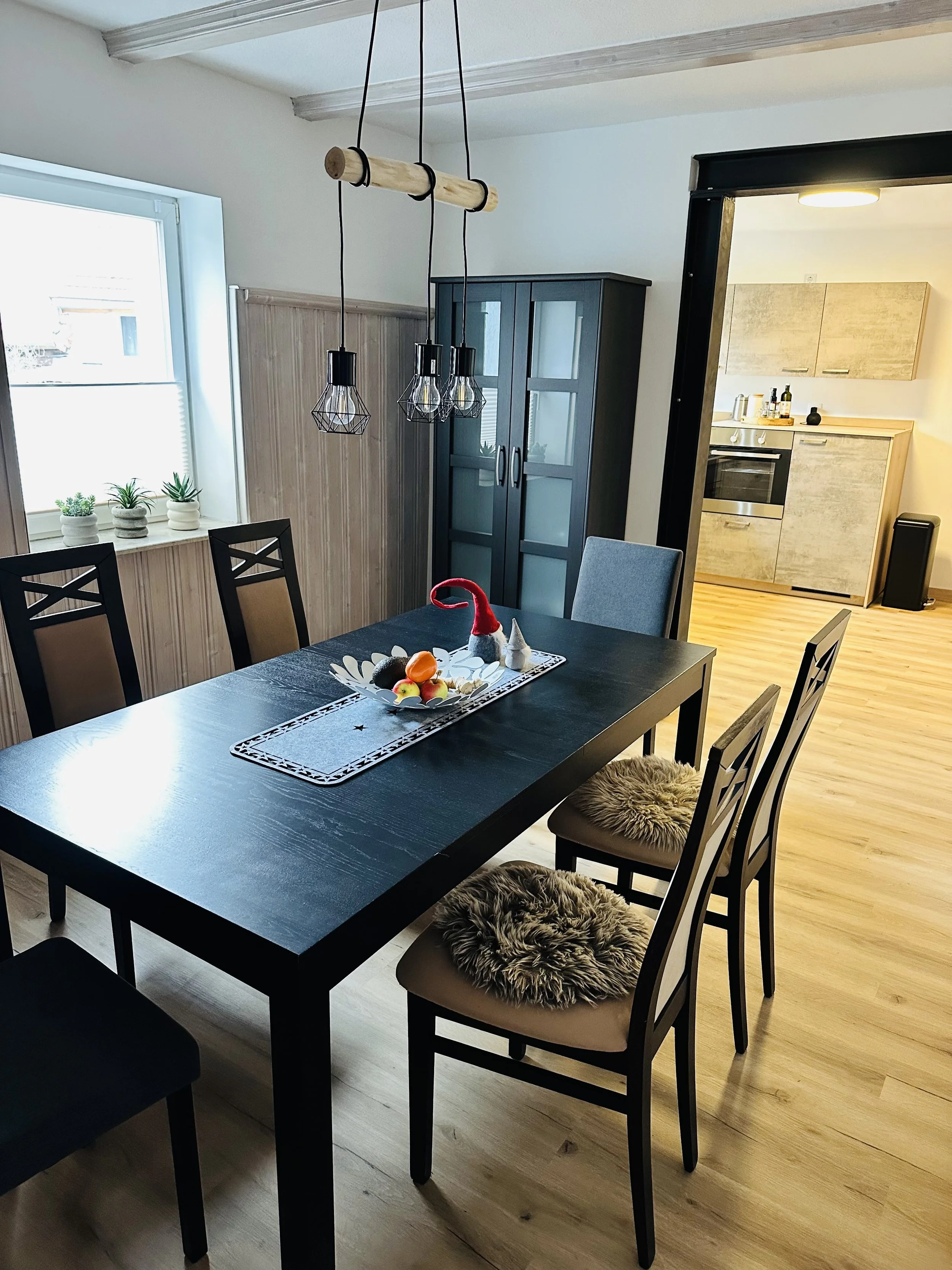 Ein Esstisch mit vier Stühlen, auf denen Fellkissen liegen, in einem Raum mit Holzboden. Im Hintergrund eine dunkle Küchenschrank und eine Fensterbank mit drei kleinen Pfl anzen. Über dem Tisch hängen drei geometrische Hängeleuchten.
