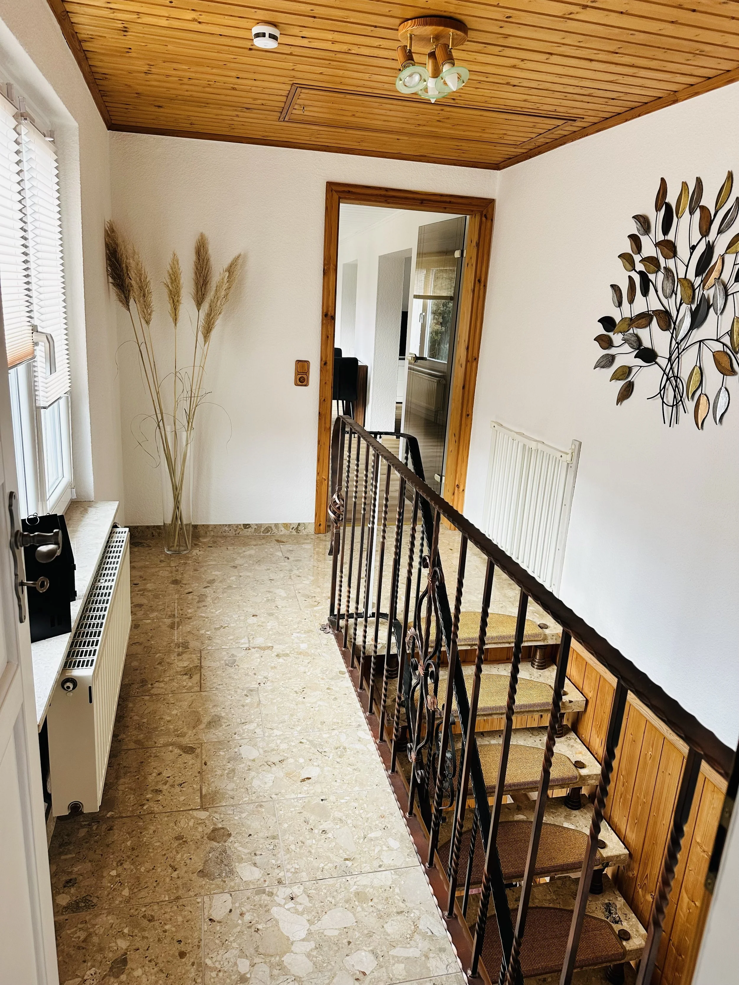 Ein Flur mit Marmorboden, einer weißen Wand, einer Holzdecke und einer Treppe mit Holzstufen und schmiedeeisernen Geländer. Es gibt einen großen Ständer mit Dekokräutern und eine Wanddekoration mit metallischen Blättern.