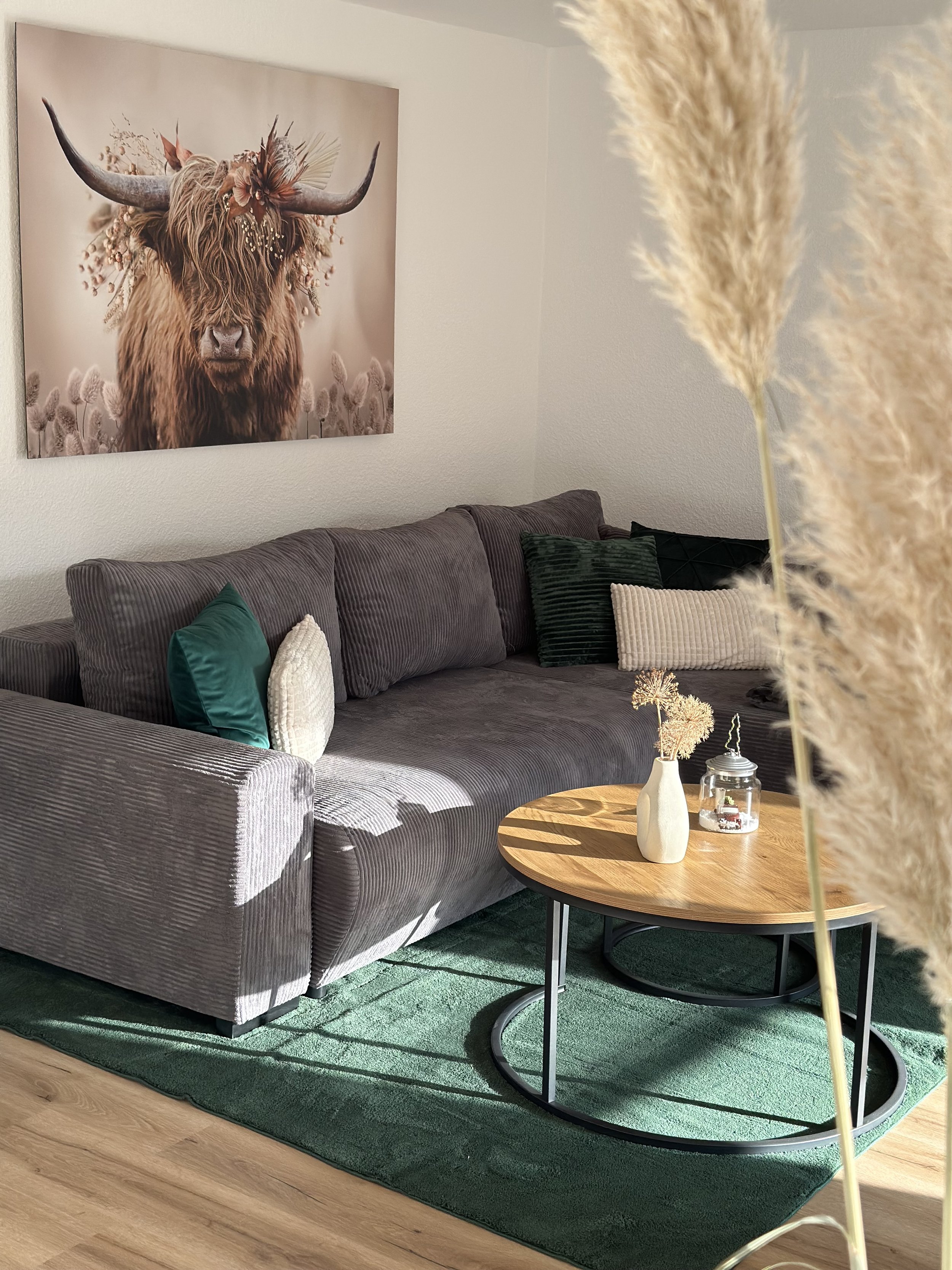 Wohnzimmer mit grauem Sofa, mehreren Kissen, Holztisch mit weißen Vasen und Kerzen, grünem Teppich, Wandbild eines Hochlandrindes mit Blumen als Kopfdeko.