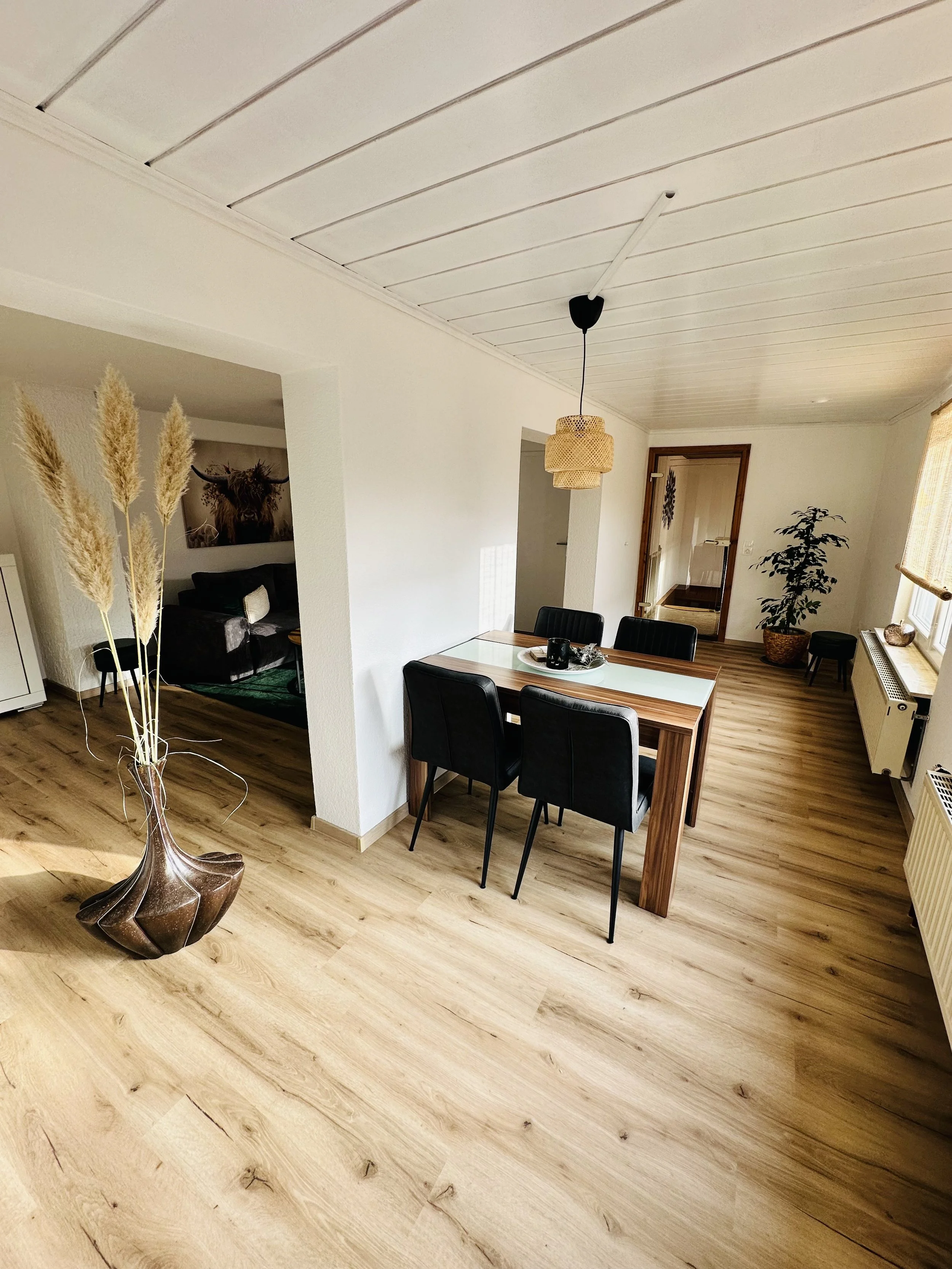 Ein Esszimmer mit einem Holztisch, sechs schwarzen Stühlen, einer Lampe an der Decke, einer Pflanze im Topf und großen Fenstern mit Rollos, die Tageslicht hereinlassen.